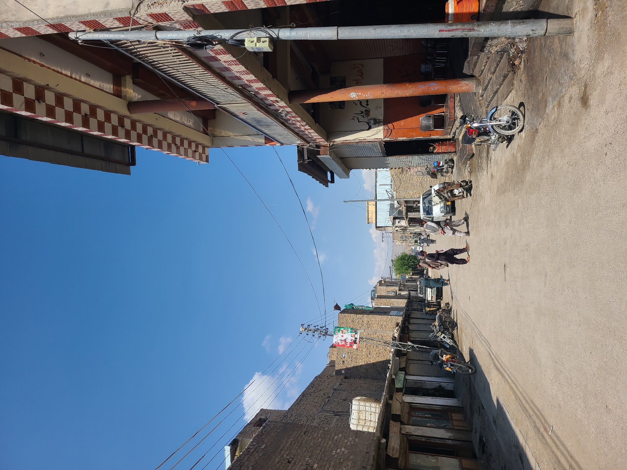 This photo shows a street in Quetta City Tehsil, Pakistan. The street is lined with buildings of varying heights and architectural styles, some with awnings or overhanging structures. Motorbikes are parked along the side of the street. Several people are walking along the road. Power lines are strung across the sky. A building displays posters featuring political or religious imagery. The sky is blue with sparse clouds.