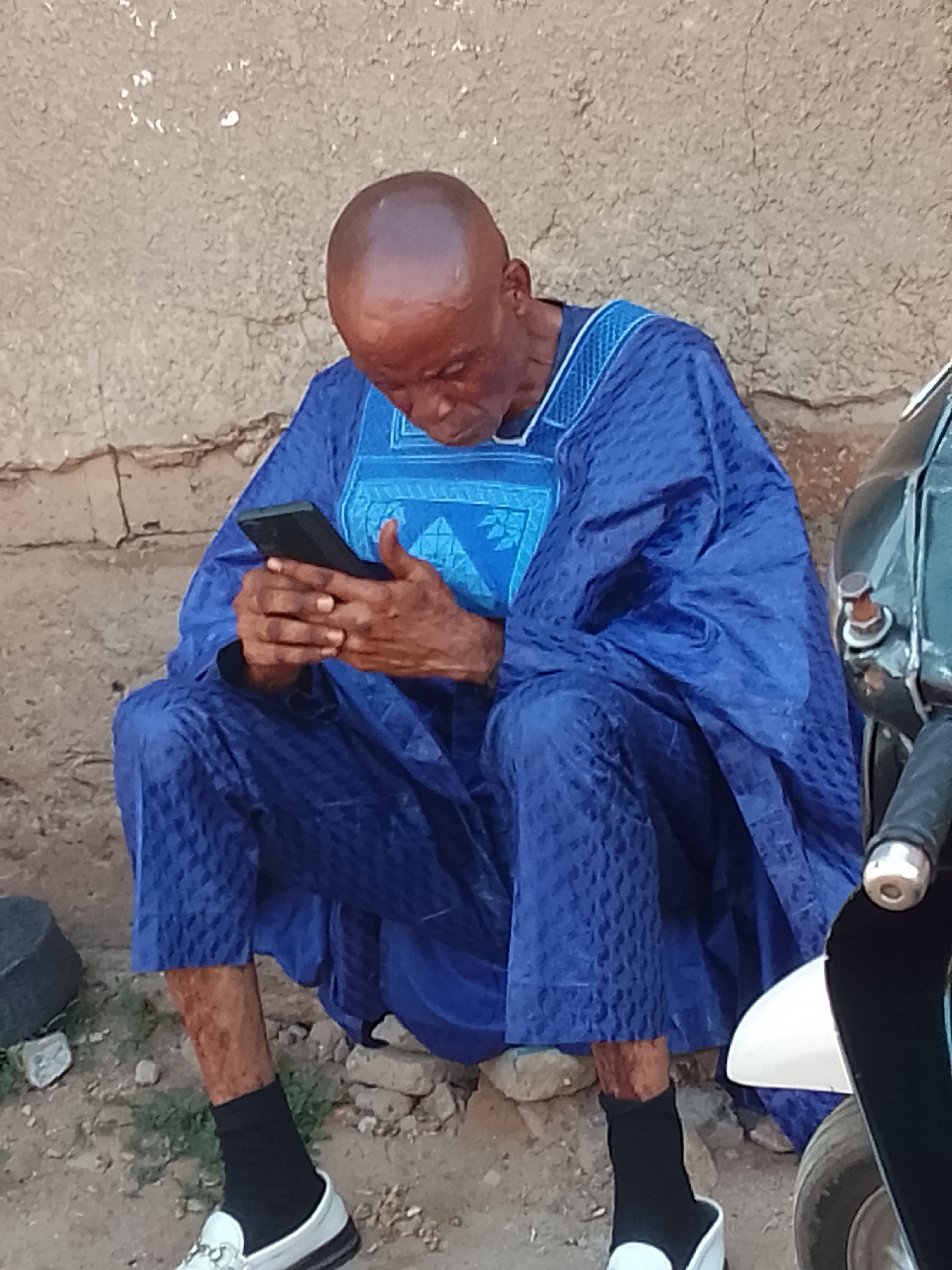 An elderly man with a shaved head, wearing a blue patterned traditional robe and pants, sits on the ground with his legs crossed. He is looking down at a black smartphone held in his hands. His left leg is visible below the robe, revealing a black sock and a white loafer. To the right of the man, a portion of a motorcycle is visible, including a white fender and a part of the handlebars. The background consists of a textured, light-colored wall and a dirt and gravel ground.