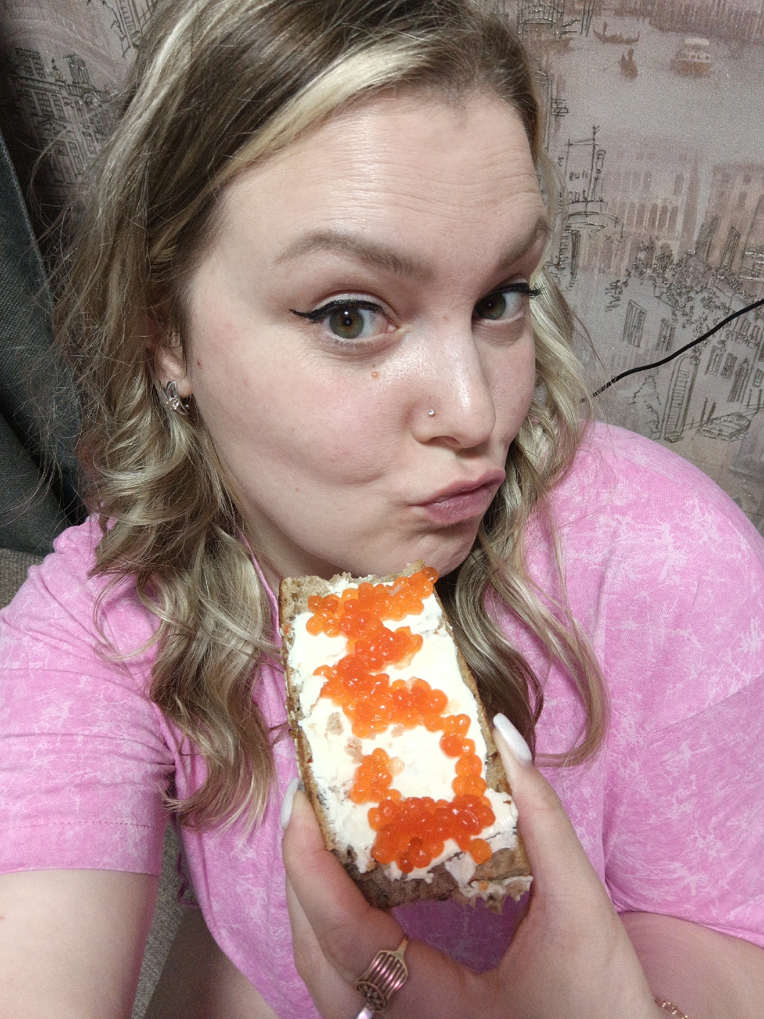 A close-up portrait features a woman holding a sandwich with red caviar and cream cheese or butter. She has light-colored, wavy hair with blonde highlights and green eyes. Her gaze is directed towards the viewer, and she is making a playful "pout" or "kiss" face. She wears a nose piercing, small earrings, and a distinctive ring on her left hand. Her nails are long and white. She is dressed in a pink t-shirt with a subtle pattern. The sandwich is made with dark bread, generously spread with a white creamy layer, and topped with a vibrant orange-red caviar. The background shows a patterned wallpaper depicting what appears to be stylized architectural elements or a city scene, possibly resembling European buildings or a landscape. The lighting suggests an indoor setting, but the time of day is not explicitly indicated. There is no visible text in the image.