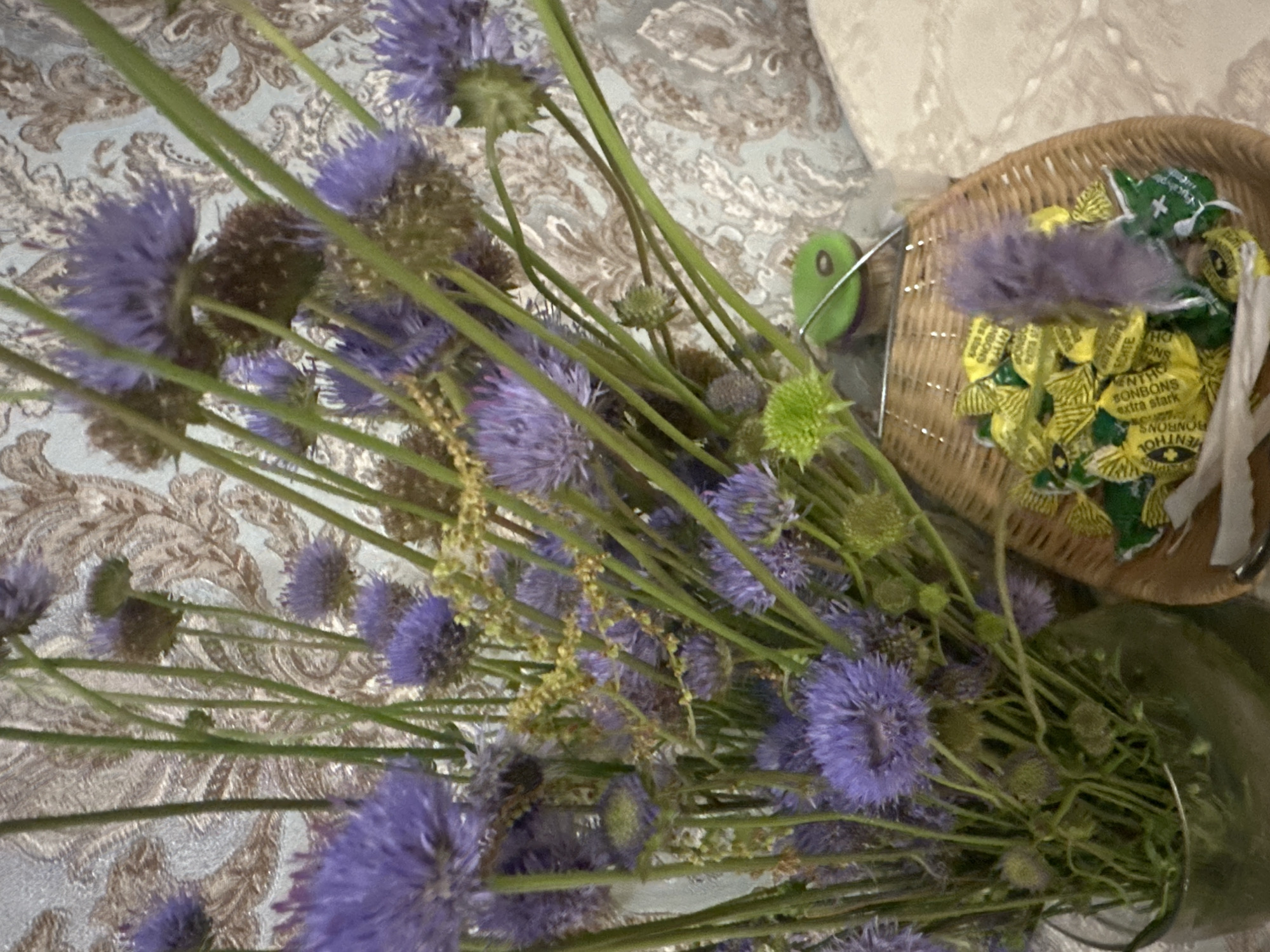 The image shows a still life arrangement of purple flowers in a vase, next to a wicker basket filled with individually wrapped yellow candies. The background is a patterned wallpaper. There is no indication of a specific city or location within the image.