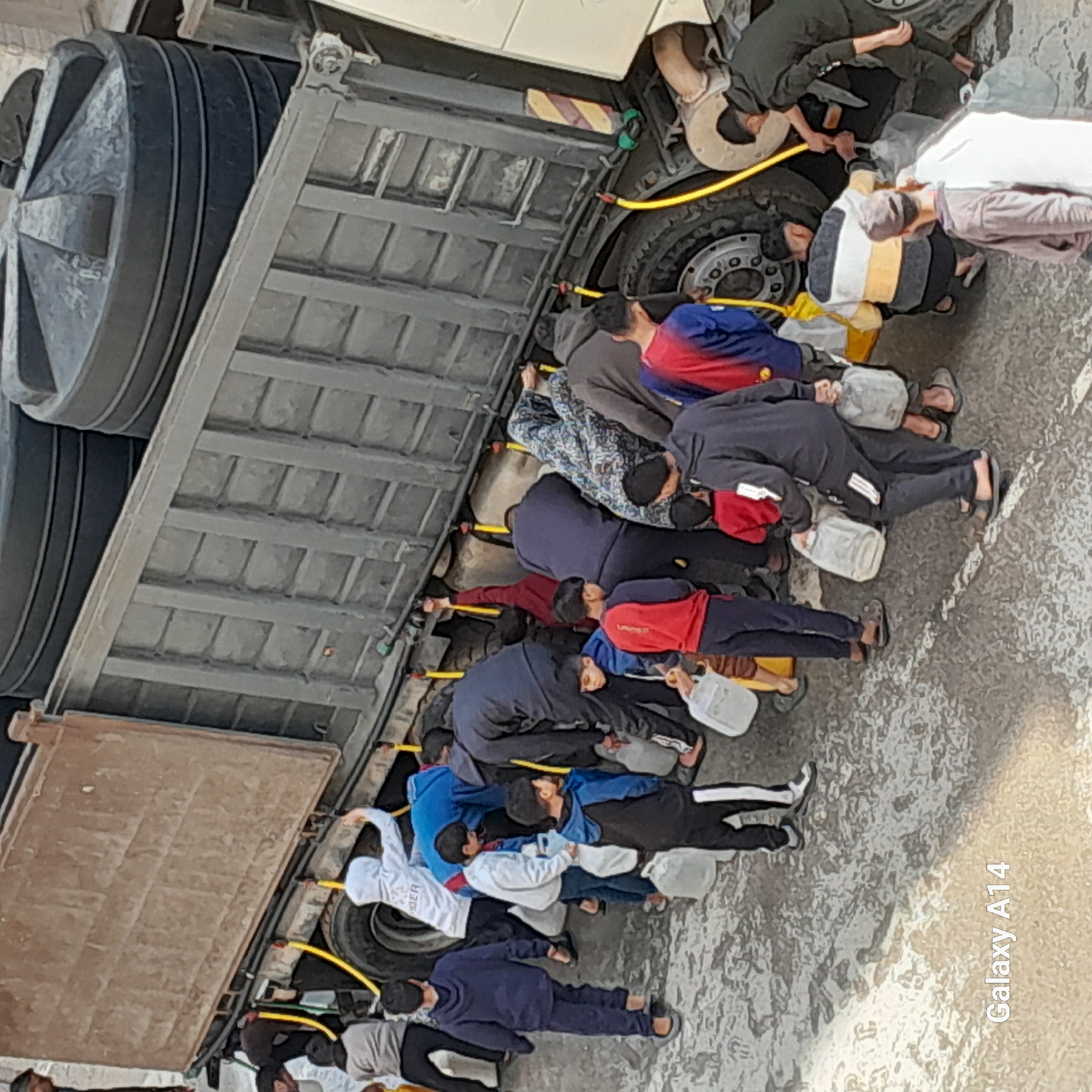 The image shows a group of people lying on the ground next to a large truck, each holding a container, possibly filling it with something from the truck via a yellow hose. The location is in a city of Unknown, Unknown.