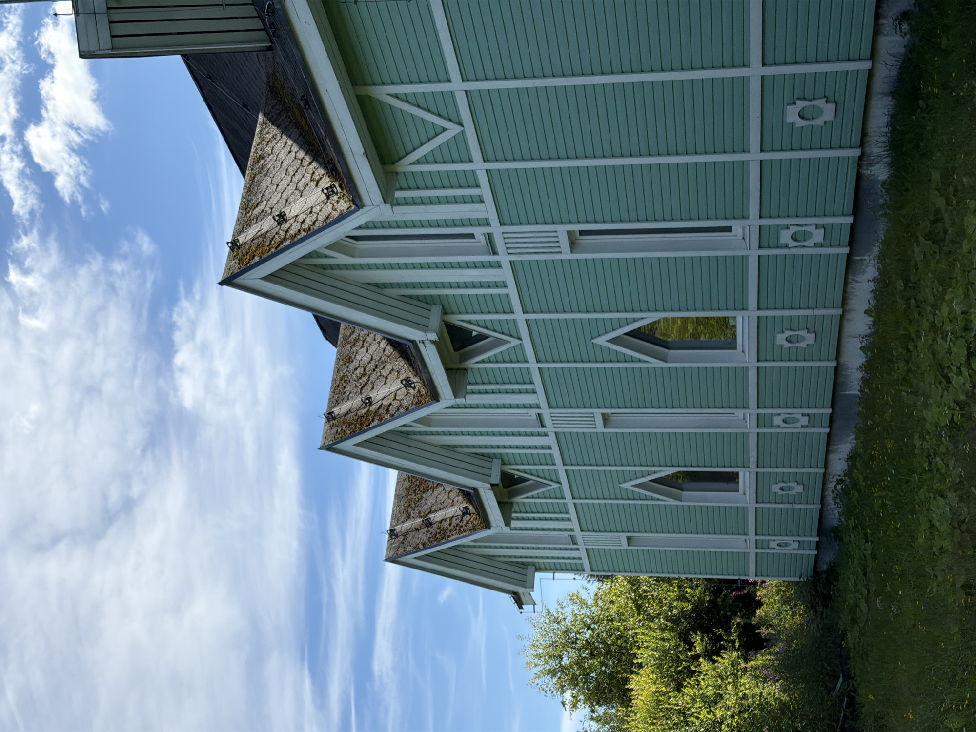 A light green, wooden building with white trim stands in an outdoor environment under a partly cloudy blue sky. The building has a multi-gabled roof covered in gray shingles that show patches of moss. Several narrow, white-framed windows are visible on the building's facade, some with pointed arch tops. The siding of the building is horizontal and painted a light green color, with white decorative elements and trim. To the left of the building, there is lush green vegetation and trees. The foreground consists of a grassy area with some smaller plants and flowers. The image appears to be taken from a slightly low angle, looking up at the building. The building is located in the city of Läskelä, Russia.