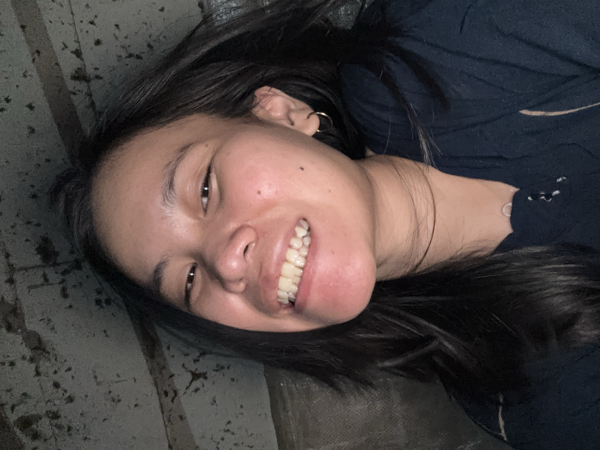 A young woman with dark hair smiles widely, revealing her teeth. She is wearing a dark blue shirt and a gold hoop earring in her left ear. The background appears to be a dimly lit, textured wall, possibly made of concrete. The image was taken in East Jakarta, Indonesia.