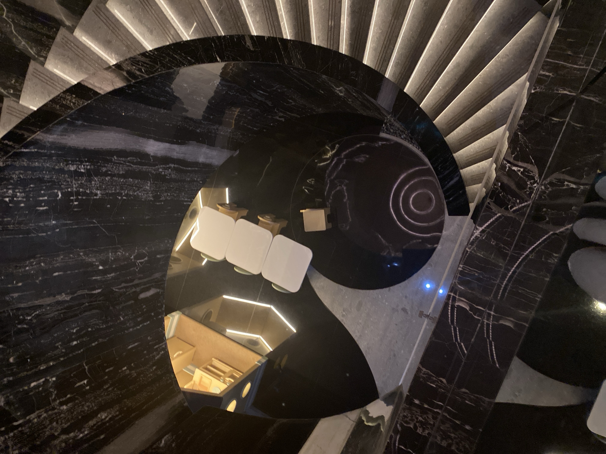 The image shows a spiral staircase with dark marble accents and illuminated steps, suggesting a modern, possibly upscale interior. At the base of the staircase, a reflective surface, likely a table or floor, mirrors the surroundings and shows a reflection of a seating area with white chairs and wooden accents. The scene appears to be indoors, possibly in a building with a contemporary design. Without further context or visual cues, it is impossible to determine the specific location in Central Jakarta, Indonesia. There are no people clearly visible in the image, though the feet of what appears to be one person are visible at the bottom of the frame, suggesting someone is standing there.