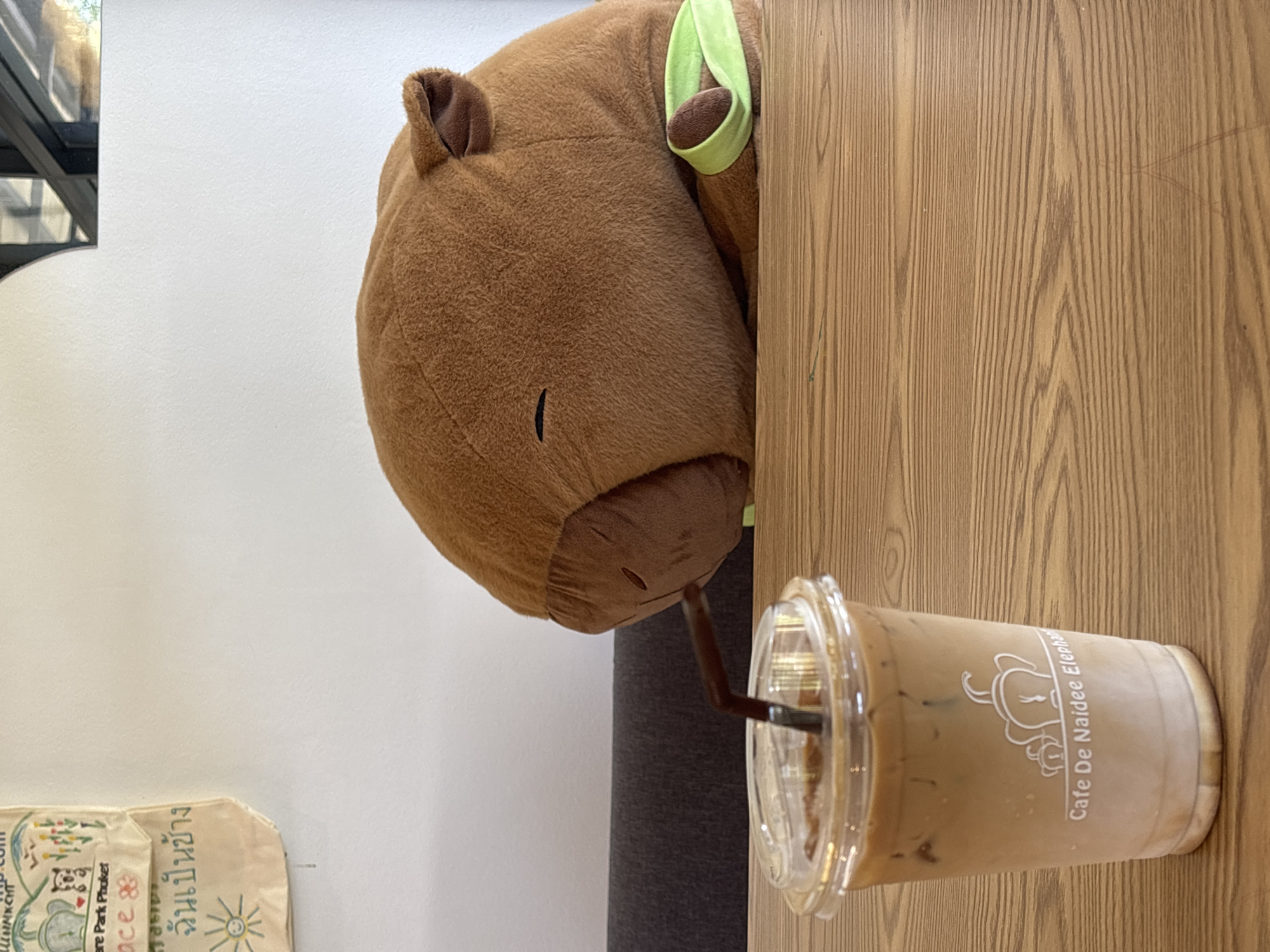 A brown plush hippopotamus is adjacent to a disposable clear plastic cup of iced coffee with a straw, on a wooden surface. The cup is labeled "Cafe De Naidee Elephant," placing the location in Choeng Thale, Thailand. The hippopotamus has a light green band around its head. A gray cylindrical object is present behind the cup and plush toy. The background consists of a white wall and a partial view of a black-framed structure. A shopping bag is in the lower left corner.