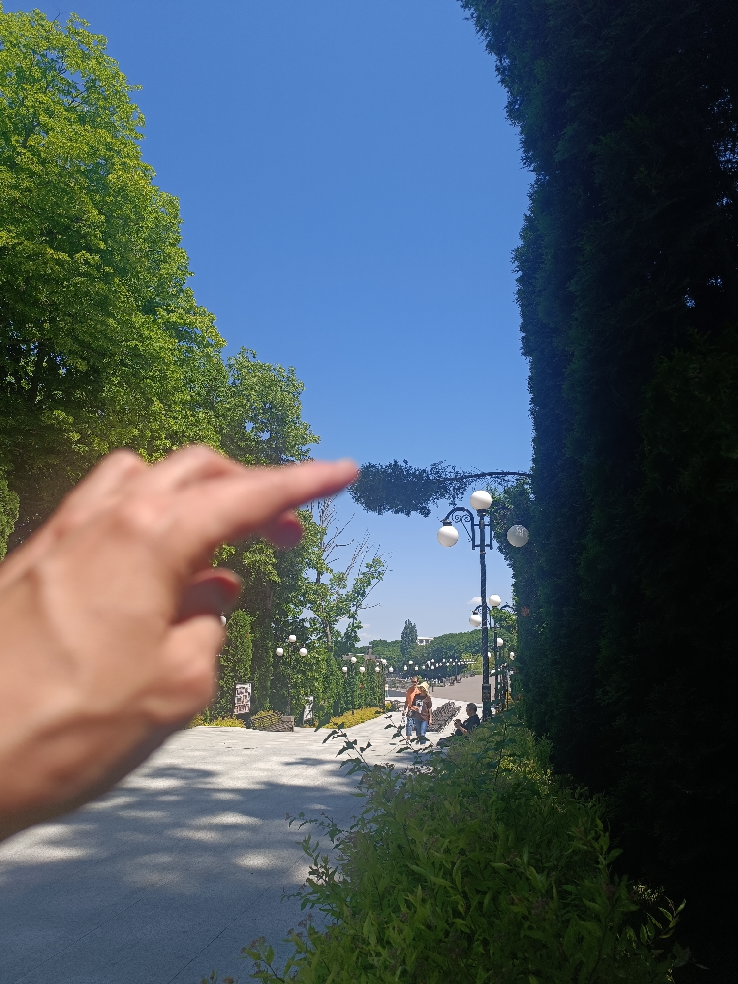An outdoor scene in Zheleznovodsk, Russia, features a pathway lined with trees and lampposts, bathed in sunlight under a clear blue sky. In the foreground, a blurred hand with an extended index finger points towards the right. Further down the paved pathway, two individuals are walking, and another person is seated on a bench. The path leads towards a distant row of trees. The environment appears to be a park or a public walkway.