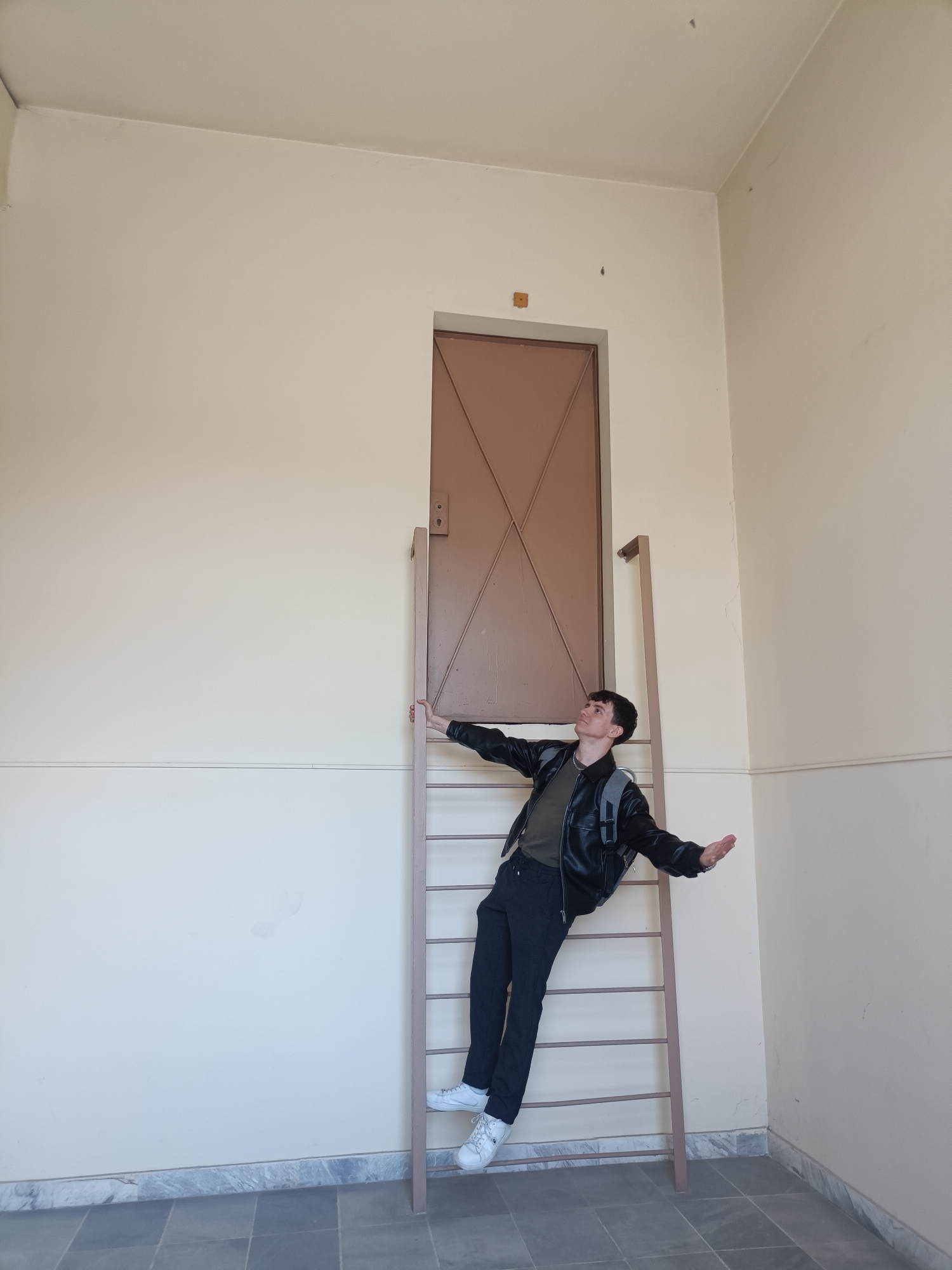 A young man is captured in a whimsical pose, standing on a ladder and leaning backward with his arms outstretched, looking upwards. He is positioned in an indoor setting, against a light-colored wall, with a tall, brown door situated unusually high above the ladder.
The man has short, dark hair and is wearing a dark green t-shirt, a black leather-like jacket, black pants, and white sneakers. A grey backpack is visible on his shoulders. His posture suggests a playful or theatrical gesture, as if defying gravity or marveling at something above.
The ladder he stands on is a simple, metal structure painted a brownish-beige color, featuring several rungs. It is placed directly beneath the brown door, which extends nearly to the ceiling. The door itself has a distinctive criss-cross pattern across its surface and a visible door handle or lock mechanism on its left side. Above the door frame, a small, square wooden block is affixed to the wall.
The walls are a uniform light cream or beige color, with a darker beige horizontal stripe running along the lower section, just above the floor. The floor is composed of large, dark grey square tiles, bordered by a grey and white marbled baseboard. The lighting is bright and even, indicating an indoor environment, possibly a hallway or large room. There is no visible text in the image.