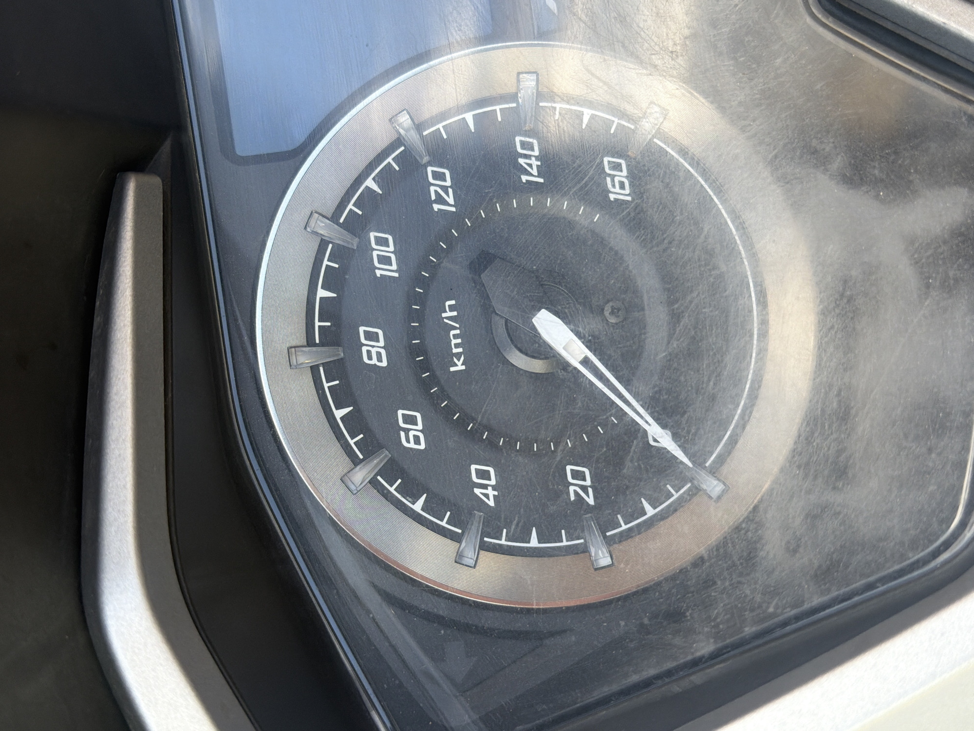 The image shows a close-up of a dusty motorcycle speedometer. The speedometer has markings for speed in kilometers per hour, ranging from 20 to 160. The needle is pointing between 40 and 60, closer to 40. The speedometer is set within a metallic bezel and the surrounding area appears to be part of the motorcycle's dashboard, with a dark, textured plastic finish. The background is a blurred expanse of a light blue, possibly sky or a reflective surface, with faint hints of what could be foliage or trees. There are no people visible in the image. The scene is indoors or in a shaded area, as indicated by the shadows cast on the dashboard. This image was captured in Baan Chaweng, Thailand, specifically in a vehicle.
