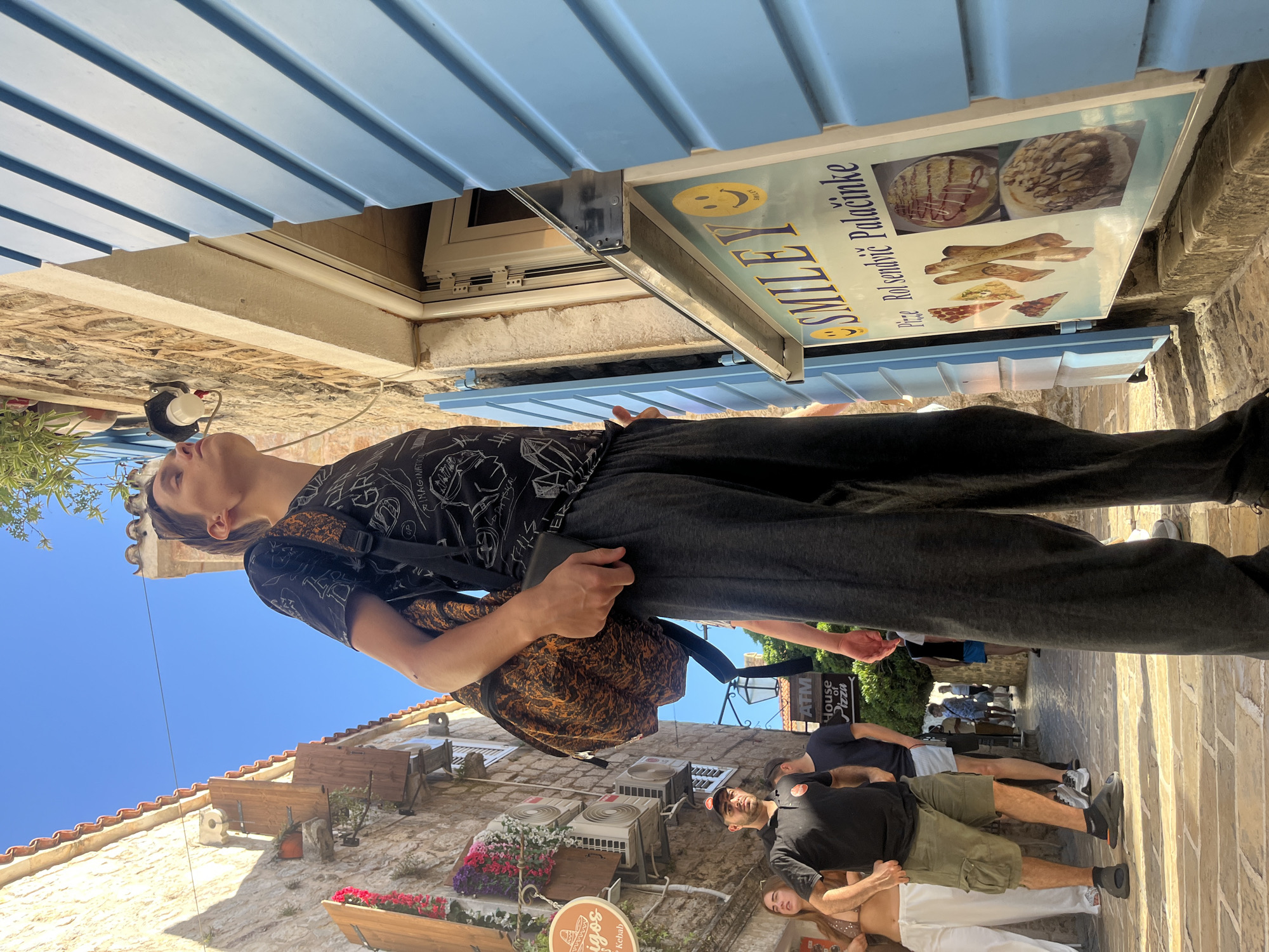 The image depicts a sunlit street scene in Budva, Montenegro, captured from a low angle looking upwards. In the foreground, a young person with dark hair, wearing a dark, patterned short-sleeved shirt, dark trousers, and a backpack, stands with their head tilted upwards, looking towards the bright, clear blue sky. A partial word, "IMAGINATIVE," is visible on their shirt.
Above and to the right of the main subject, a prominent sign for "SMILEY" is displayed, featuring a yellow smiley face logo. The sign advertises "Rosendijć Palačinke" (pancakes/crepes) and "Pizza," with accompanying food illustrations. A blue awning or shutter system extends across the upper part of the image, partially covering the building where the sign is mounted.
The street is paved with light-colored stone, and the surrounding buildings are constructed from traditional stone, some with terracotta roof tiles visible in the distance. Flower boxes filled with red and purple flowers adorn the facades, and several air conditioning units are mounted on a wall in the mid-ground.
Several other individuals are present in the background, casually walking or standing on the street. Among them, a man with a beard and a black cap is visible. Further down the street, additional commercial signs can be seen, including "ATM House of Pizza" and a circular sign displaying "Amigos" and "Kebab." The strong sunlight and clear shadows indicate a bright, sunny day, likely midday or early afternoon. The scene conveys a lively, pedestrian-friendly atmosphere in an old town setting.