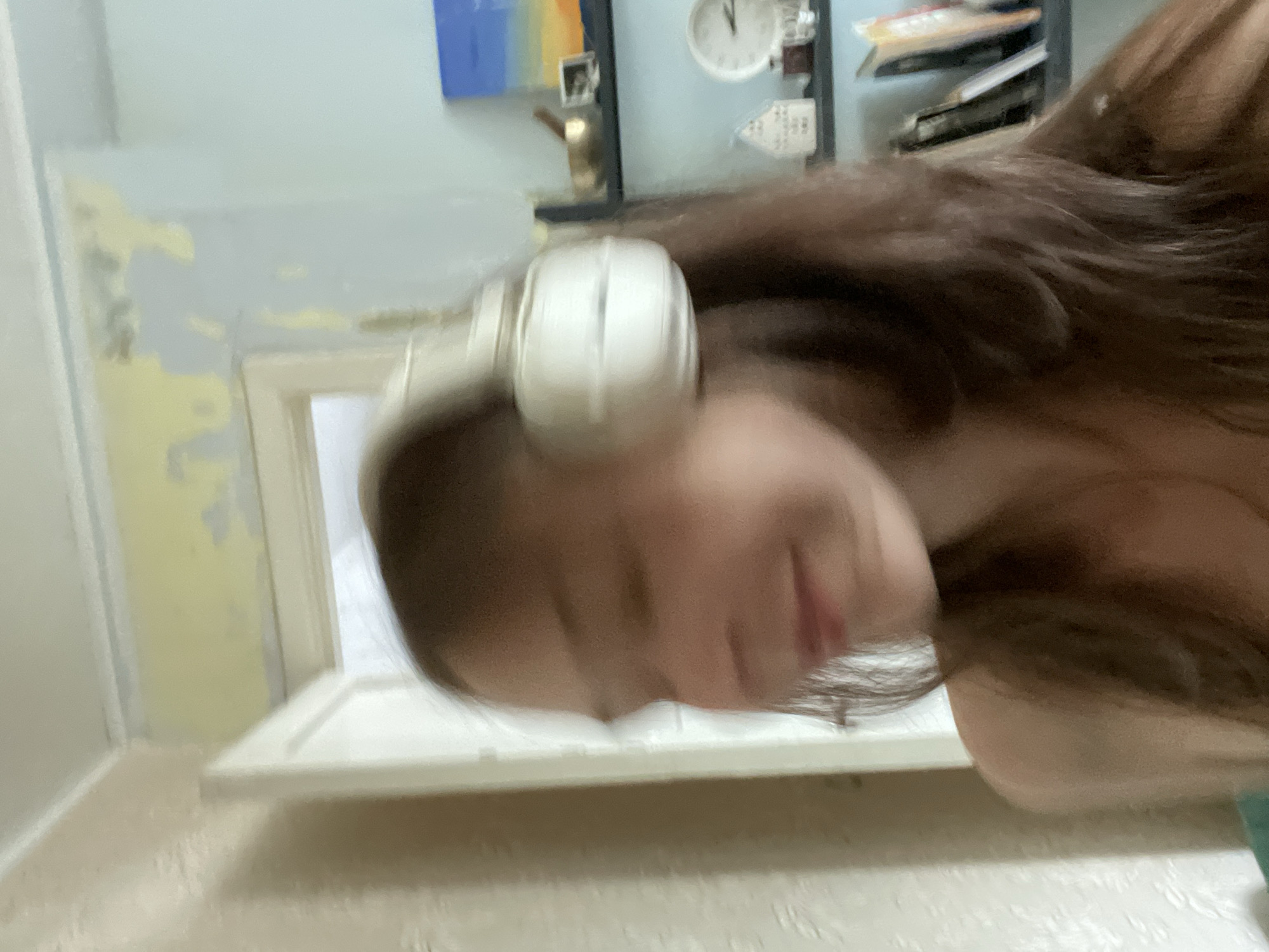 The image shows a young person with long brown hair wearing white over-ear headphones. They are looking to their left and smiling. The person is in a room with light blue and pale yellow walls. There are shelves in the background with books, a clock, and a small painting. The image appears to be a selfie taken by the person themselves.