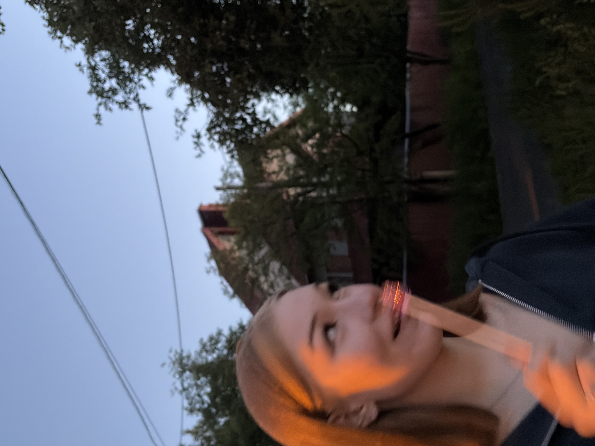 The image captures a young woman in Новинки, Russia, during what appears to be late afternoon or early evening. She is positioned in the left foreground, with her left side illuminated by a strong, warm orange light. She has light-colored hair, a faint smile, and her gaze is directed upwards and to her right. She wears a dark, zippered garment, possibly a hoodie, and a silver necklace is visible around her neck. Her right hand holds a translucent stick with a glowing red tip near her mouth. The background is out of focus, showing a clear blue sky with two prominent parallel power lines. Below the power lines, the blurred forms of several trees with dark foliage and residential buildings with red roofs are discernible, one featuring a visible chimney. The lower right foreground reveals a dark path or road with a lighter line and overgrown dark green vegetation.