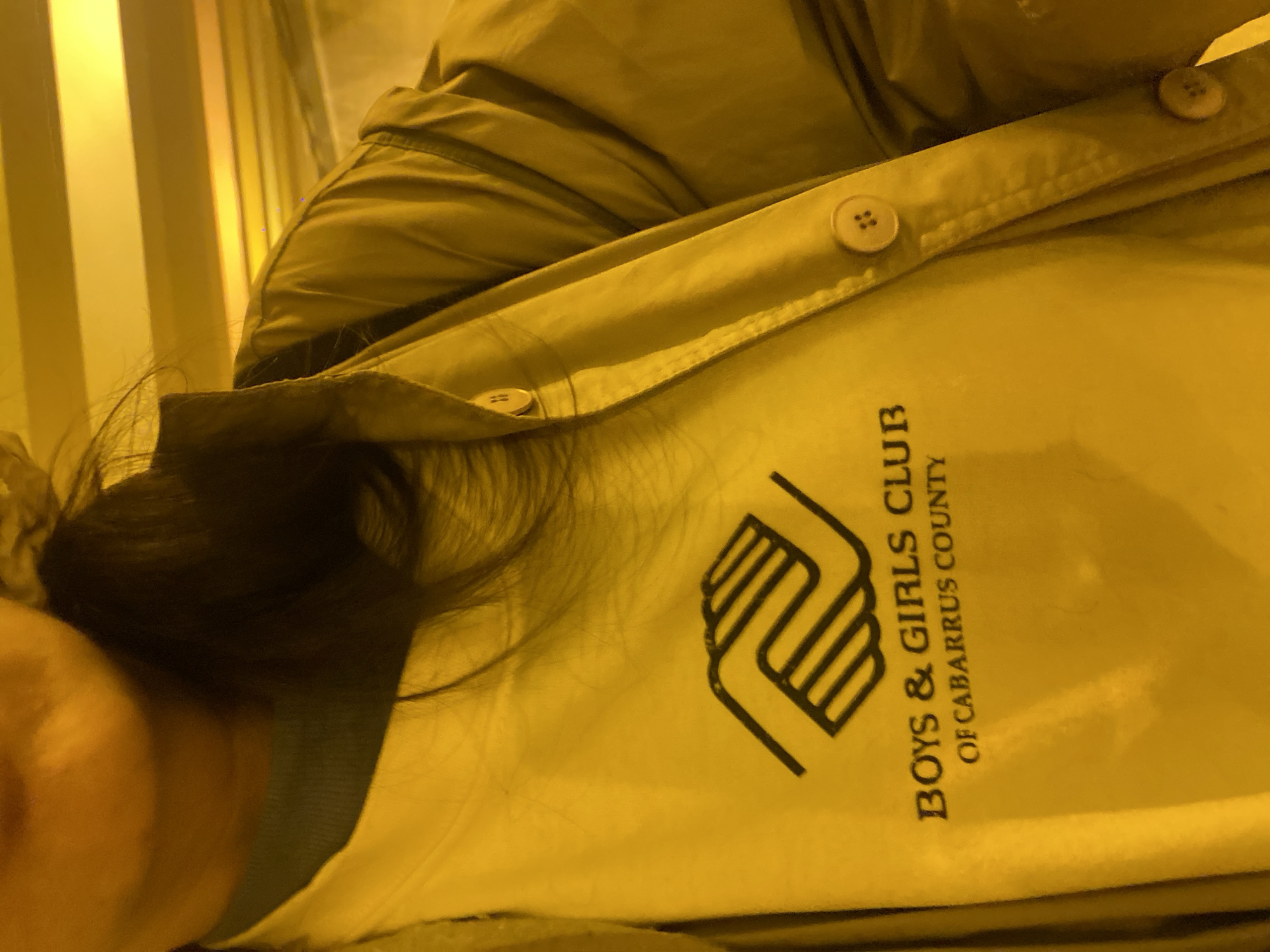 A close-up, high-angle portrait captures the upper body of an individual, likely female, under intense yellow illumination. The subject is wearing a light-colored, long-sleeved shirt displaying the "Boys & Girls Club of Cabarrus County" logo in black, layered over a green neck garment, and partially covered by an unbuttoned, light-beige collared jacket with prominent circular buttons. The individual's lower face, including the mouth with closed lips, is visible. The background is dominated by distinct horizontal bands of strong yellow light and shadows, characteristic of architectural lighting found within an underground transit system. Based on the distinctive yellow lighting and structural elements, the scene is situated within the Moscow Metro system in Moscow, Russia.