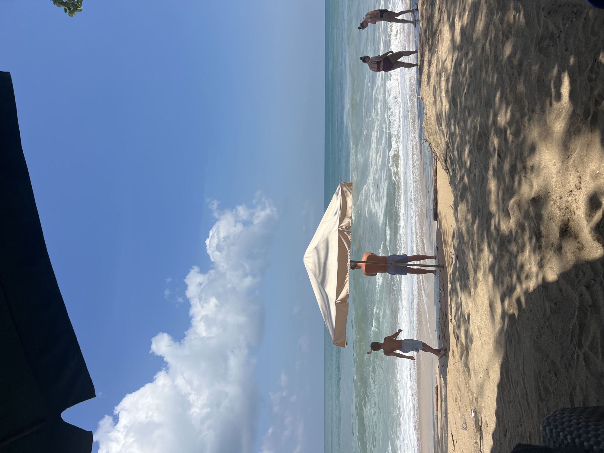 A beach scene in Kuta, Indonesia, under a clear blue sky with scattered clouds. The foreground is sandy with dappled shadows from an overhead structure, possibly an umbrella or awning. In the middle ground, a large, light-colored umbrella stands on the beach, providing shade. To the left of the umbrella, a young boy wearing light-colored shorts is walking towards the water. To the right of the umbrella, a man wearing blue striped shorts is standing. Further to the right, near the waterline, two women are standing. One woman wears a dark red bikini, and the other wears a dark bikini. The ocean is calm with gentle waves breaking on the shore. The horizon is visible, with the sea meeting the sky.