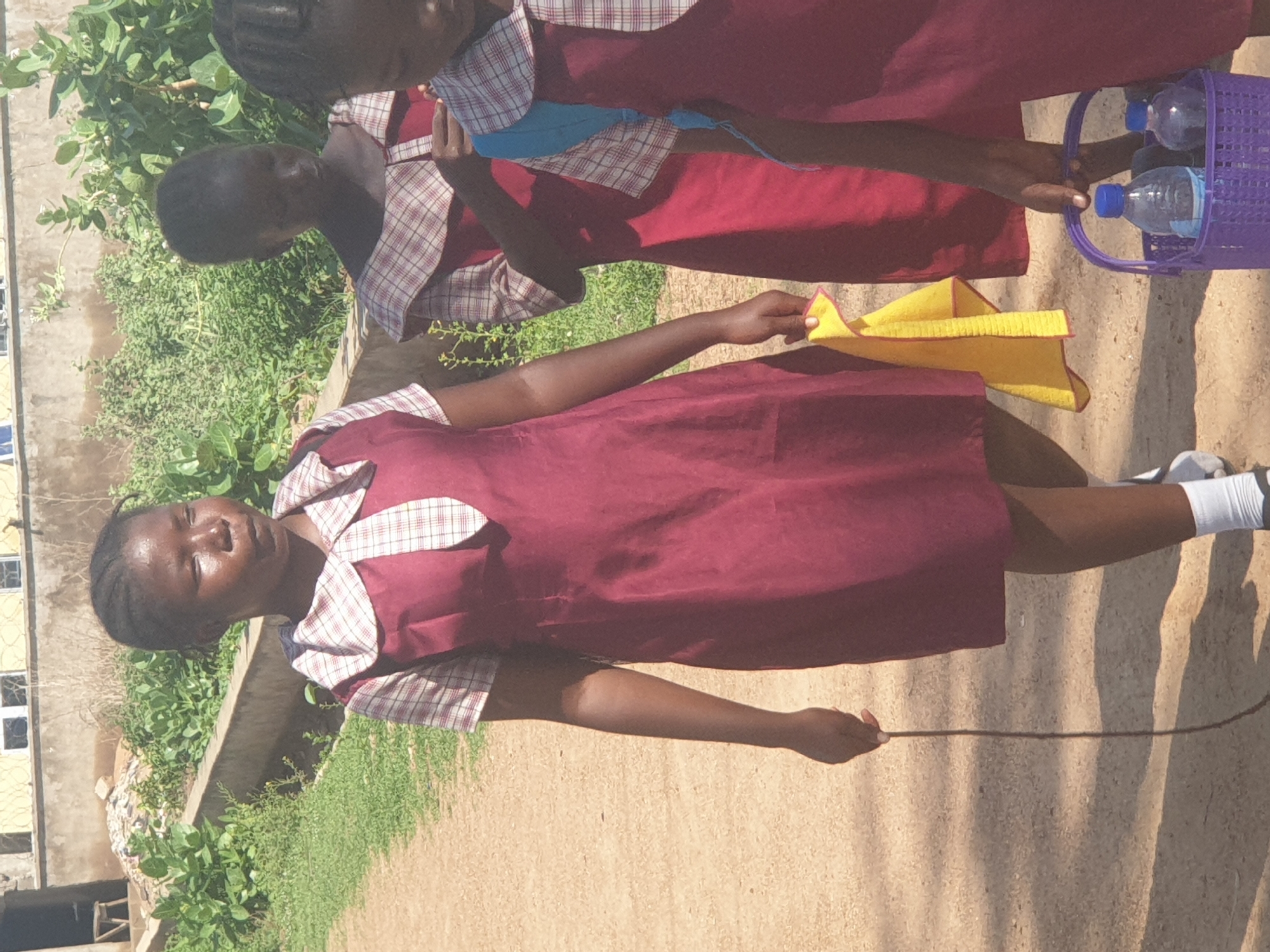 In Jalingo, Nigeria, three young girls in maroon school uniforms with plaid collars walk along a dirt path. The girl in the foreground, on the left, holds a thin stick in her right hand and a yellow cloth in her left. She wears white socks and dark shoes. To her right, another girl in a similar uniform walks with her head down. A third girl, partially visible on the far right, also wears the school uniform and has a blue backpack strap visible. Behind the girls, a low concrete structure runs along the path, with lush green vegetation growing beyond it. A building with windows is visible in the background.