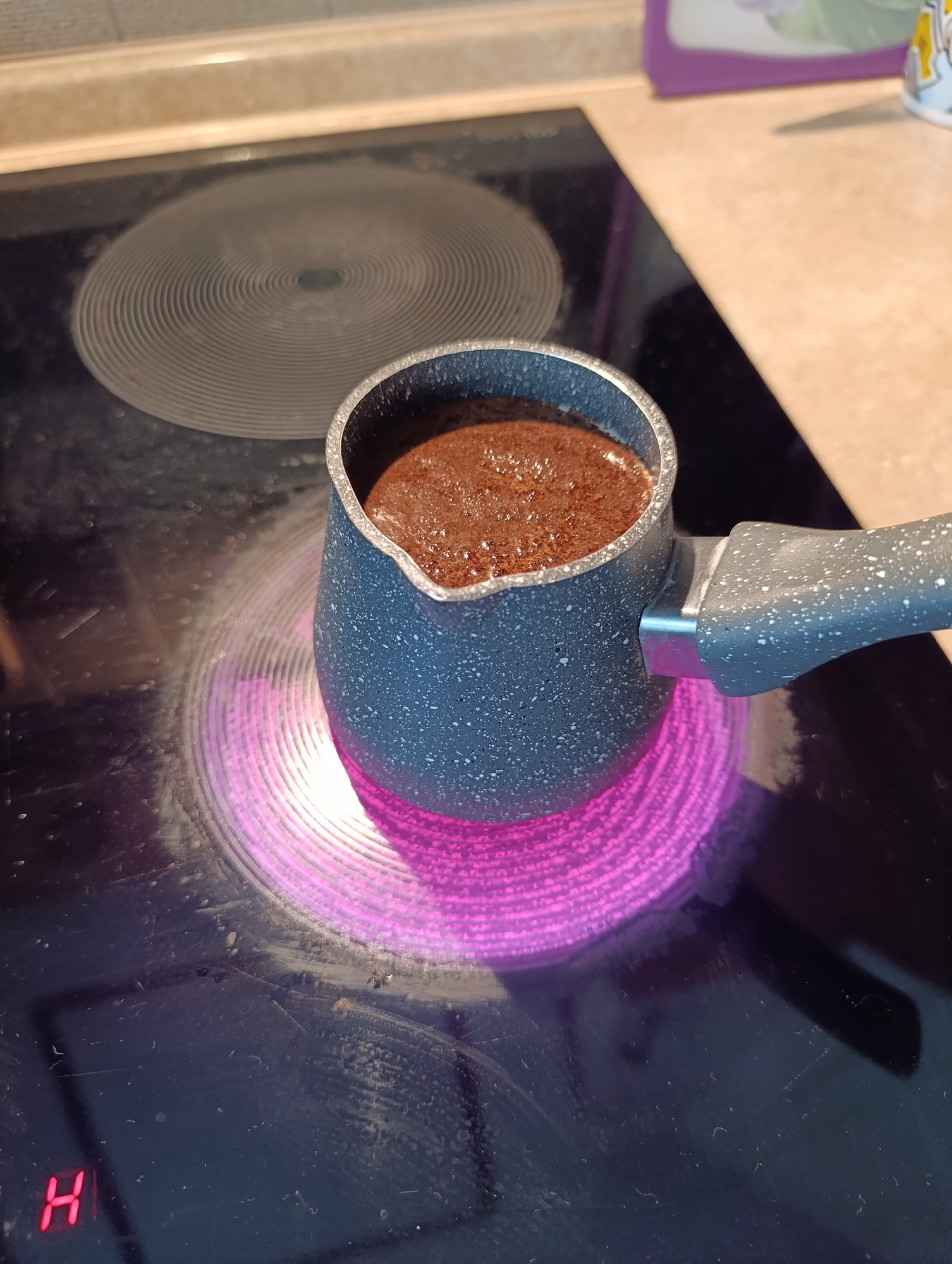 A cezve filled with coffee is heating on an electric stovetop. The stovetop burner is glowing purple, indicating it is on a high setting. The coffee appears to be brewing and is starting to foam. The background shows a countertop and some kitchen appliances. The scene is indoors and appears to be in a kitchen. There is no visible indication of the location being in Moscow, Russia.