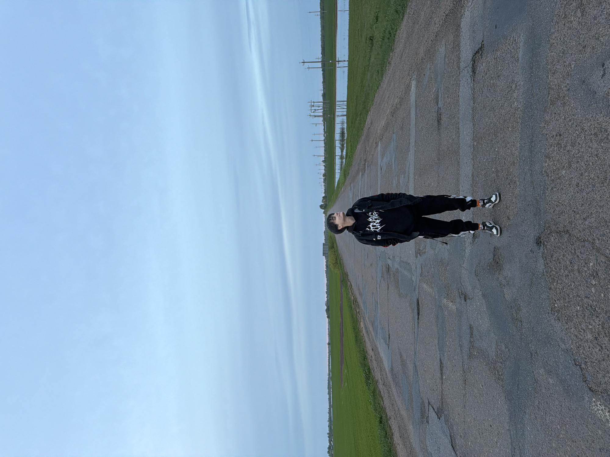 A person stands on a paved road, adjacent to a grassy field that appears to be flooded. Overhead power lines are visible in the distance. The sky is overcast. The person is wearing a black jacket with a white graphic, black pants with white stripes, and sneakers. The setting is Azyakul', Russia.