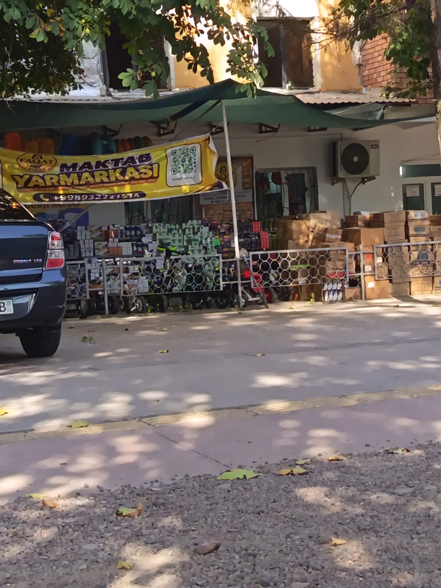 This is a medium-shot, eye-level, outdoor photograph of a street scene in Angren, Uzbekistan. A dark colored car is parked on the left side of the frame, with its rear bumper and taillight visible. In the center of the image, a store front displays a large yellow banner that reads "MAKTAB YARMARKASI" in large, bold letters, along with a phone number that begins with "+998932227574" and a QR code. The banner is partially obscured by the overhanging branches of a tree with green leaves and some yellow fruit. Behind the banner, the store interior is visible, showing shelves stocked with various items. In front of the store, several racks of merchandise, possibly school supplies or household goods, are arranged on the sidewalk. These racks are filled with boxes and containers of different sizes and colors. Cardboard boxes are also stacked along the exterior wall of the building, to the right of the merchandise racks. An air conditioning unit is mounted on the white wall of the building above the store entrance. The ground in front of the store is paved with concrete and gravel, with scattered fallen leaves. The overall scene suggests a commercial area on a sunny day.