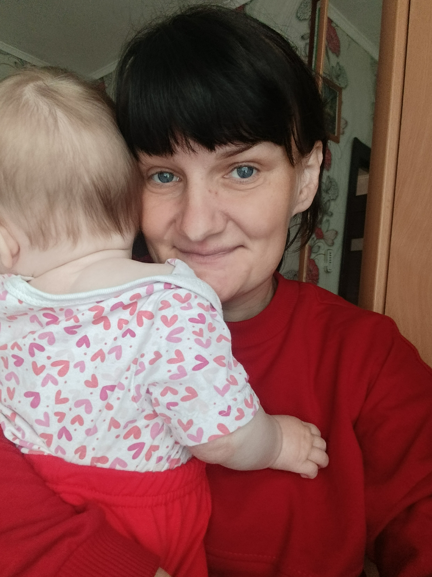 A woman with short black hair and blue eyes is holding a baby. The baby has light hair and is wearing a white shirt with pink hearts and red pants. The woman is wearing a red sweater. Behind the woman is a wall with floral wallpaper. A door is visible to the right of the woman. The location may be Фоки, Russia, based on provided information.