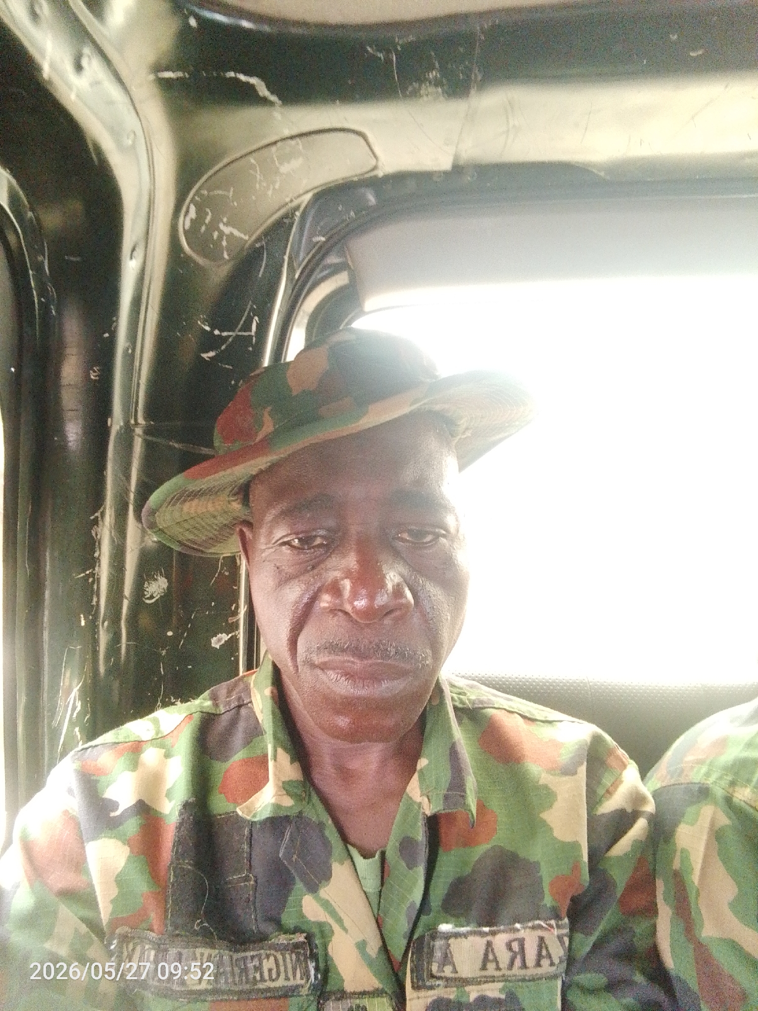 A middle-aged man in a camouflage uniform and hat sits inside what appears to be a vehicle. The interior of the vehicle is dark and metallic. The man's uniform has patches with text, one of which appears to say "NIGER" and the other "ARMY," possibly indicating he is a soldier or military personnel. The image is taken indoors, likely inside a vehicle in the city of Lokoja, Nigeria, and is time-stamped 2026/05/27 09:52.