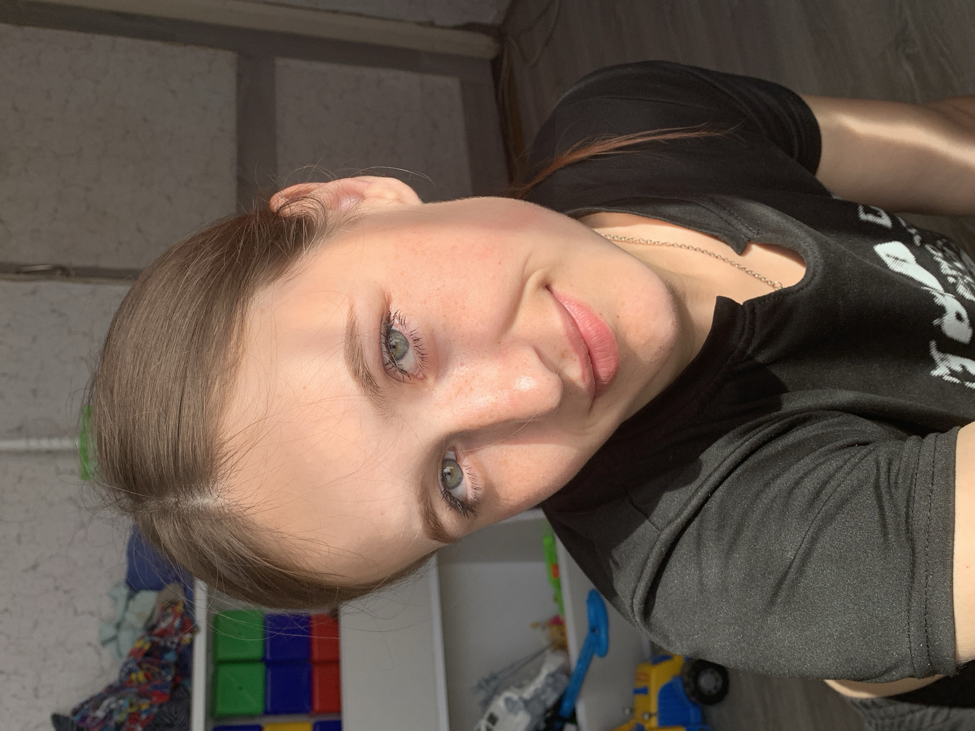 A fair-skinned young woman with freckles is looking at the camera. She has brown hair pulled back from her face and light-colored eyes. She is wearing a black, long-sleeved shirt. A silver necklace is visible on her neck. In the background, there's a child's shelf with colorful blocks and toys, as well as light coming through a window. The image appears to have been taken indoors. There is no verifiable evidence of location to indicate Фоки, Russia.