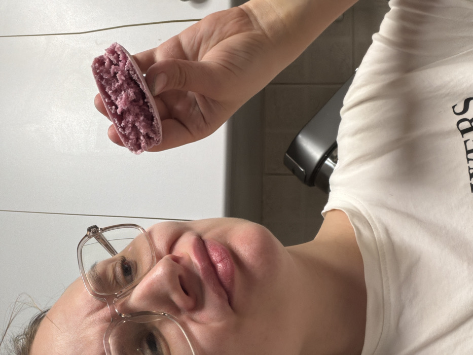The image displays a head and upper torso shot of an individual, likely female, indoors, presumably in a kitchen. The person, positioned slightly to the left and looking towards the right, wears clear-framed eyeglasses and a white short-sleeved t-shirt with partial dark lettering ("WERS") visible on the chest. Their facial expression features a slightly scrunched nose, downturned lips, and furrowed brow, suggesting a reaction of skepticism or mild displeasure. The individual holds a half-eaten or broken light purple macaron in their right hand. The macaron's exterior shell is smooth and light purple, while the exposed filling is a darker, granular purple. In the background, white or light-colored cabinetry is visible above a light-colored tiled backsplash. A dark gray or black kitchen appliance, possibly a stand mixer, is present in the lower right background. The specific city location of Tel-Aviv, Israel, cannot be determined from the visual evidence within the image.