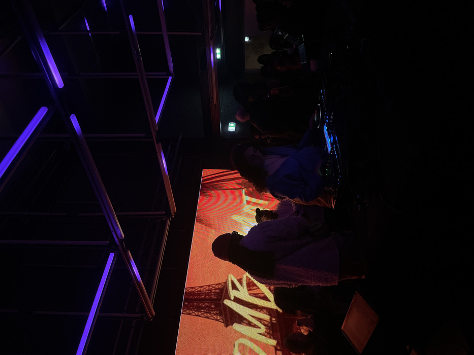 A dimly lit scene captures a group of people seemingly enjoying an event. An orange screen displaying the Eiffel Tower and the word "HOME" acts as a backdrop. Adjacent to the screen, a geometric metal structure with neon purple lighting adds a futuristic touch. People are seen standing and some are holding what appears to be cameras or phones, possibly taking photos or videos. The overall atmosphere appears to be lively and engaging.