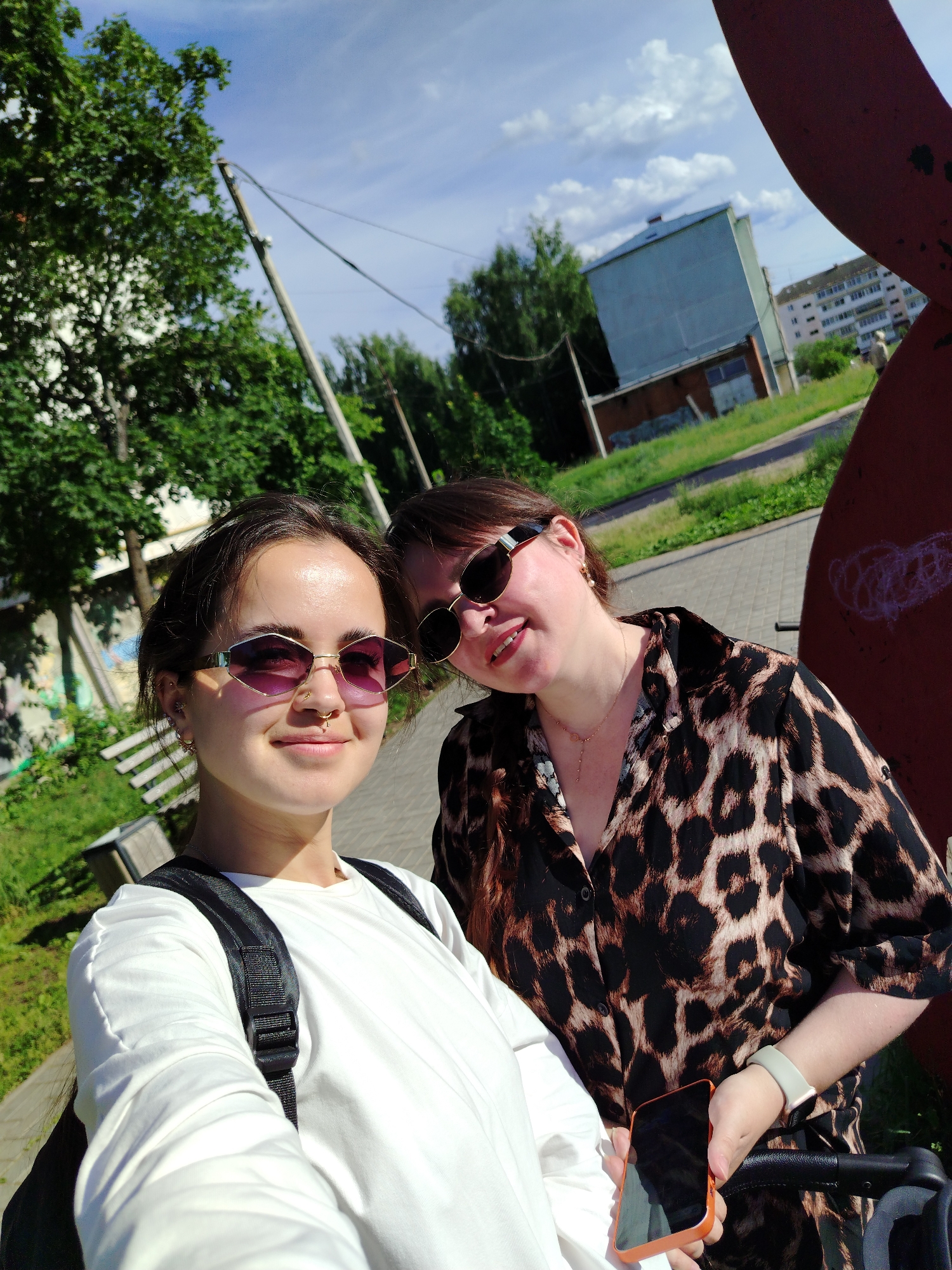 Two young women are taking a selfie outdoors on a sunny day. The woman in the foreground, on the left, has dark hair pulled back and is wearing sunglasses with purple lenses and a silver nose ring. She has on a white long-sleeved shirt and a black backpack. The woman next to her, on the right, also wears sunglasses and a leopard-print collared shirt. She is smiling and holding an orange-cased smartphone.
They are in a park-like setting with lush green trees behind them. In the background, there are buildings, a road, and a utility pole. The sky is blue with scattered white clouds, suggesting it is daytime. The overall mood appears cheerful and casual. There is no visible text in the image.