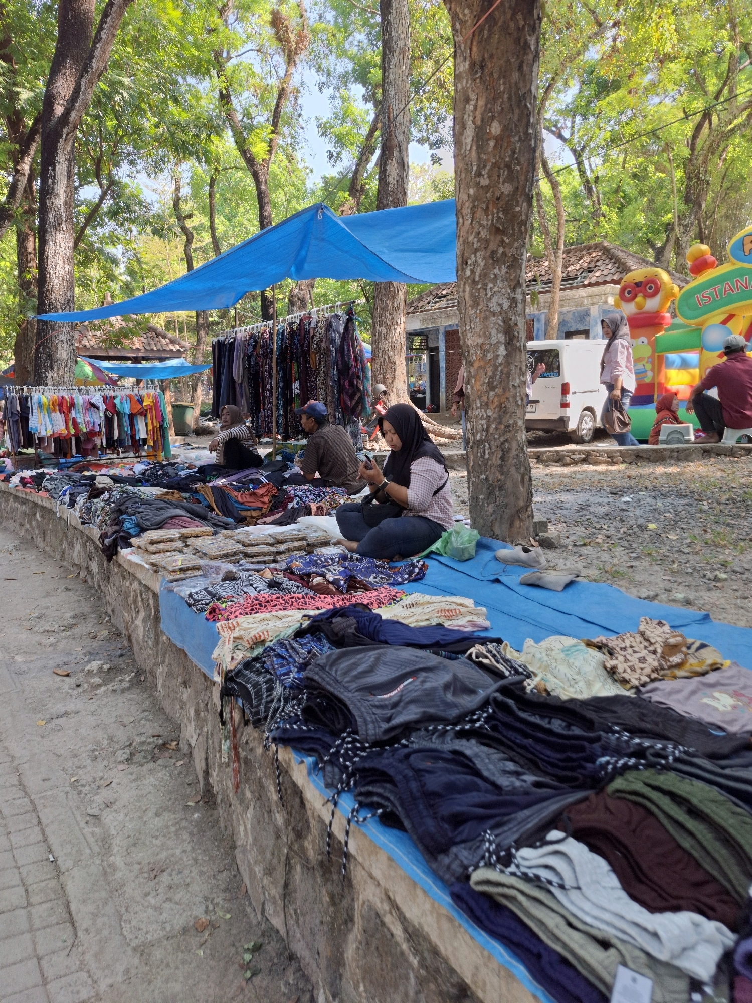 In Tawun, Indonesia, a market scene unfolds under a bright blue canopy, shaded by large trees. A diverse array of clothing items is displayed on a raised stone platform covered with blue tarps, featuring racks of neatly hung garments and piles of folded apparel. Several individuals are present, including a woman in a black hijab sitting on the ground and looking at her phone, another woman in a light pink hijab and white shirt standing near a white van, and a man in a brown shirt and blue cap looking towards the right. In the background, a vibrant inflatable playground with characters and the text "ISTANA" is visible, suggesting a family-friendly atmosphere. The ground is a mix of dirt and paved areas, with fallen leaves scattered around, indicating a natural and open-air setting.