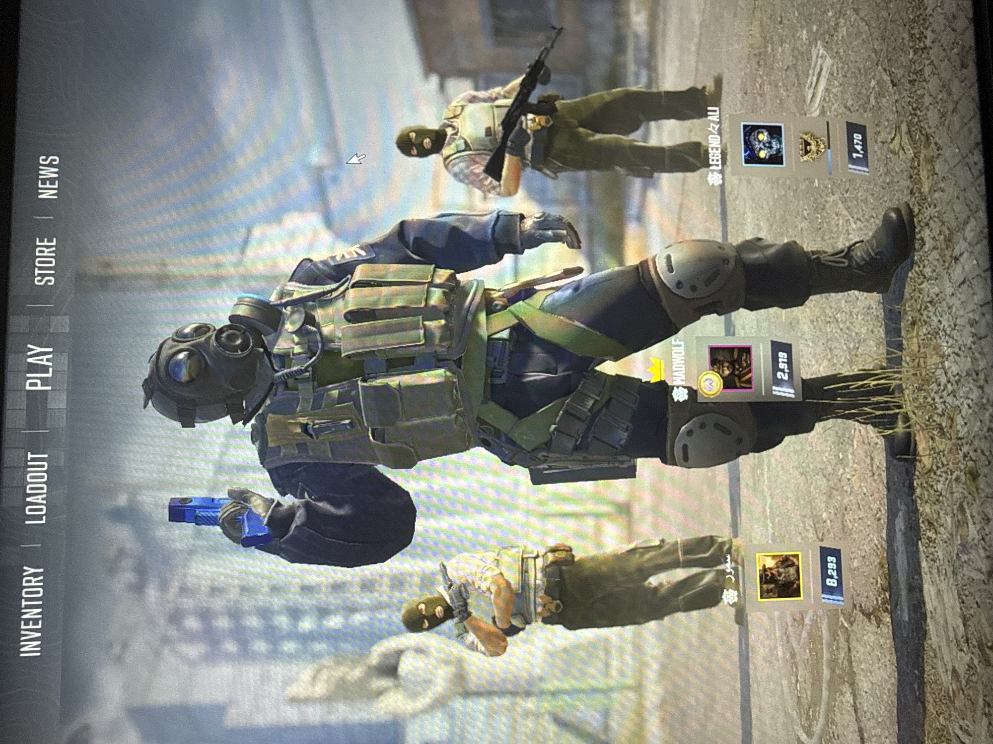 The image shows a screen display of what appears to be a video game. On the left side, there are menu options, reading "INVENTORY", "LOADOUT", "PLAY", "STORE", and "NEWS". In the center, three characters are displayed. The character in the middle is wearing a gas mask and combat gear, holding a blue handgun. Above this character is another character holding an assault rifle. The third character is below the middle character, with all three having a user name with profile information listed next to them. The profile information includes a portrait and numerical value. The background is blurry but appears to be an urban environment.
There is no evidence in the provided image that indicates the scene is in Jiulongpo District, China.