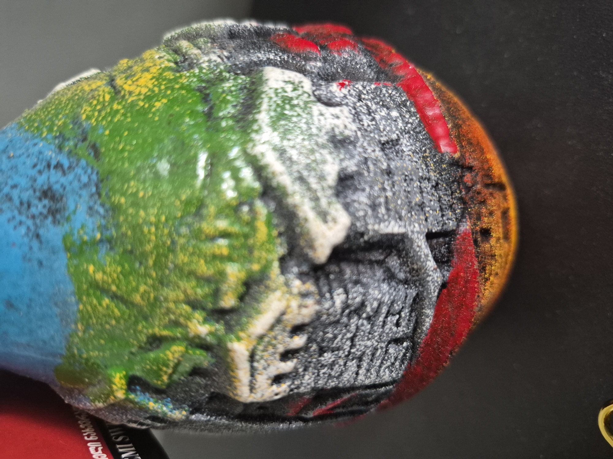 The image displays a close-up, macro-style view of a textured, irregularly spherical object. This object, possibly a bath bomb or a decorative item, features a vibrant, multi-colored surface with a distinct rough and granular texture.
The object is painted in several sections: the left side is a bright blue, followed by a vibrant green section heavily speckled with yellow dots. Adjacent to this is a textured area of white and dark grey, which exhibits sculpted relief, giving it an uneven and rugged appearance. A bold red band traverses part of the dark grey section, leading to an area of warm orange and yellow hues on the far right, which gradually fades into a dark, possibly black, background. The textured surfaces show fine particles and small raised bumps.
Underneath the object, on the bottom left, a section of a red surface is visible, bearing white text. The legible text includes "089 631 036" and "EMI SW". In the bottom right corner of the frame, a small, golden, loop-shaped object can be faintly seen against the dark background.
No activity is taking place; the image is a static shot of the object, likely taken indoors under controlled lighting to highlight its intricate details and colors. The location context for the media file is Yerevan, Armenia.