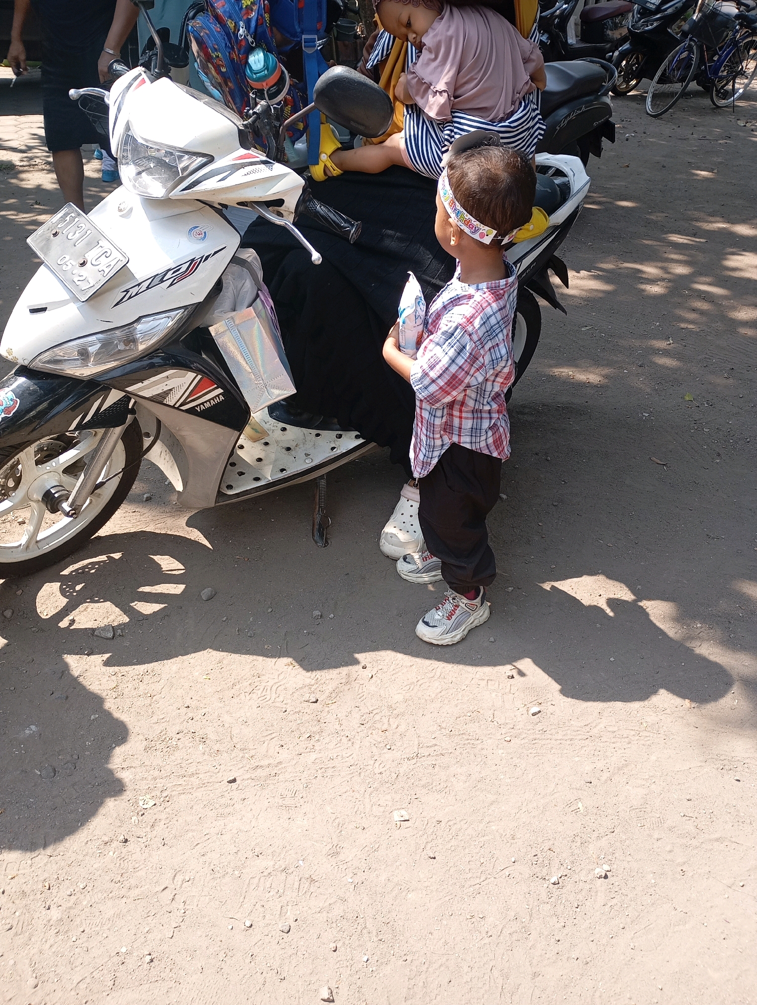 A child stands to the right of a white Yamaha motorcycle in Ketegan, Indonesia. The child, wearing a plaid shirt, black pants, and white sneakers, holds a white object. A woman, partially visible, sits on the motorcycle, holding a baby. The ground is dirt with shadows cast from trees. The motorcycle has a license plate and various stickers. Other motorcycles and a bicycle are in the background.