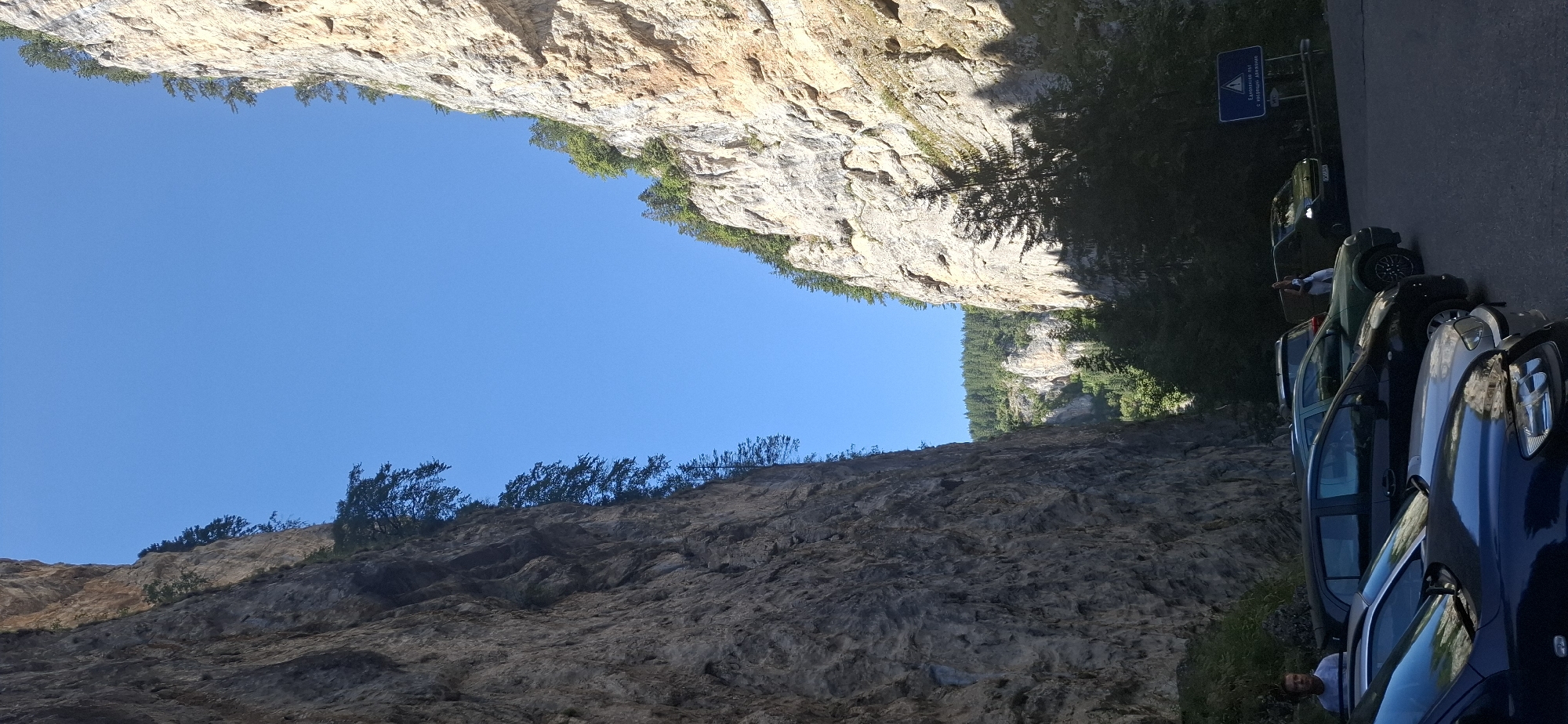 The image depicts a narrow canyon with steep, rocky cliffs on either side. The cliffs are textured and show signs of erosion, with sparse vegetation growing from crevices. Trees, predominantly conifers, are visible at the top of the cliffs and in the distance, forming a dense forest. The sky is a clear, bright blue.
In the foreground, a parking lot is visible with several cars parked. The cars are of various makes and models, including a dark blue sedan, a silver sedan, a black hatchback, and a green SUV. A white van is also present further back.
Two individuals are visible in the parking lot. One person, a man wearing a striped shirt, stands near the leftmost cars, looking away from the camera. Another person, a woman wearing a white top and dark pants, is standing near the green SUV, appearing to interact with someone inside it or looking towards the road. A blue sign with white text and a triangular warning symbol is posted near the van, indicating a potential hazard or information for drivers.
The environment suggests an outdoor recreational area, possibly a tourist destination, given the parking facilities and the dramatic natural scenery. The overall impression is a scene of natural grandeur juxtaposed with human activity in the form of a parking area and vehicles. The location is identified as the Trigrad Gorge, near the village of Trigrad, Bulgaria.
