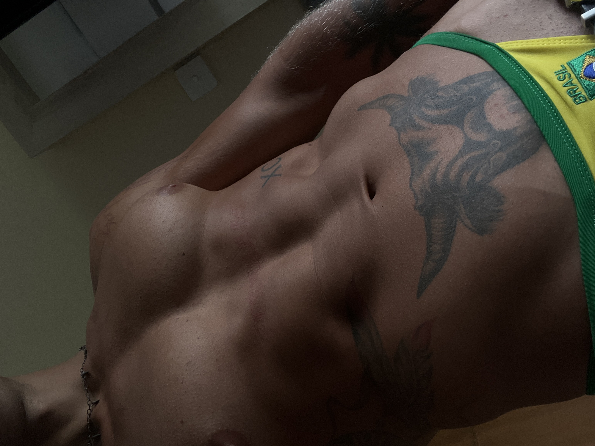 A close-up, waist-up shot of a man's torso is captured in a dimly lit room. He is wearing yellow and green briefs with the word "BRASIL" visible. The man has several tattoos, including one on his abdomen that appears to be of a bull's head, and another on his chest with the letters "XO". He is also wearing a silver chain necklace. The background is out of focus, but suggests an indoor environment with natural light coming from the right. The scene is devoid of any specific landmarks indicating a location in North Kuta, Indonesia.