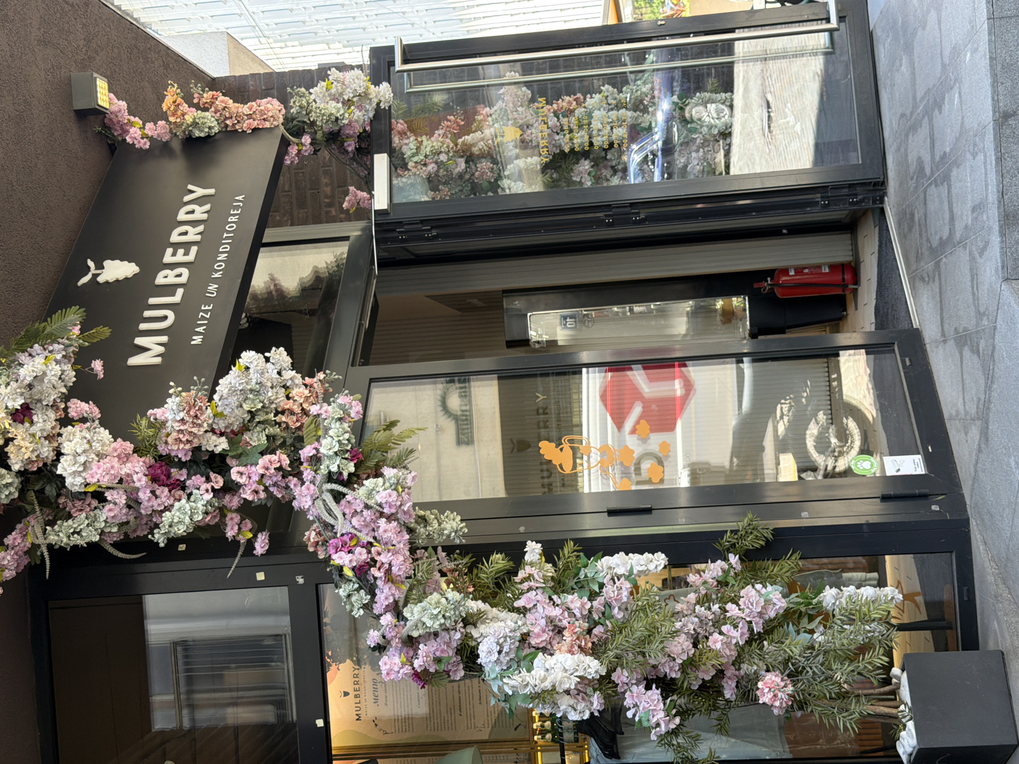 This image shows the exterior entrance to a bakery or cafe named "MULBERRY MAIZE UN KONDIITOREJA" in Riga, Latvia. The entrance features large glass doors and windows, decorated extensively with artificial pink, white, and green flowers and foliage arching over the doorway and cascading down the sides. A black sign with "MULBERRY" and a stylized white berry graphic is mounted above the entrance.
Inside the bakery, visible through the glass, are shelves, a counter, and what appears to be a menu displayed on the left. A red fire extinguisher stands near the entrance on the right. A motorcycle is partially visible inside the premises on the left. The building's facade appears to be brick and dark metal. The ground in front of the entrance is paved with grey tiles. The overall impression is of a charming and floral-themed establishment.