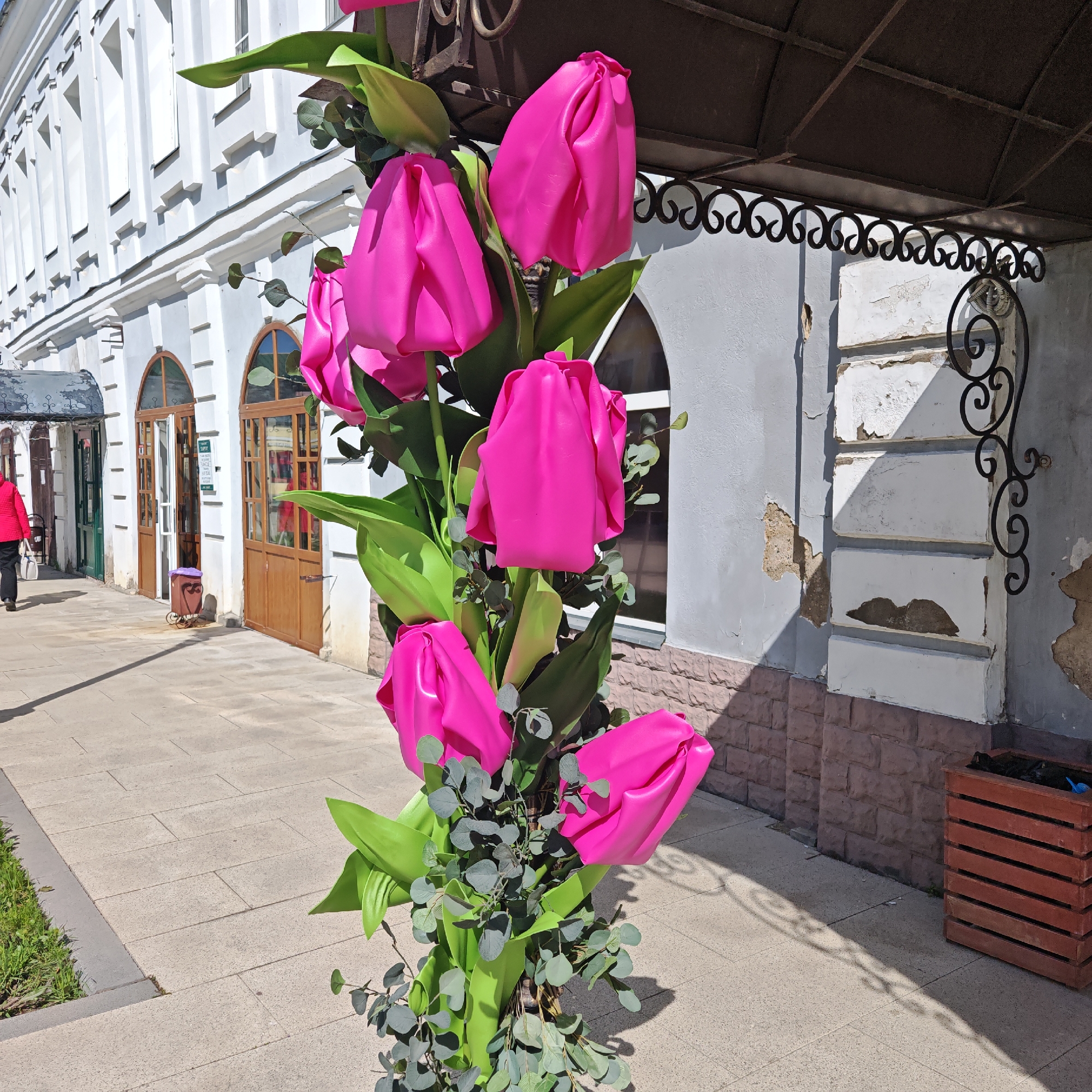 The image presents an outdoor urban scene in Rostov, Russia, featuring a large, vibrant display of artificial hot pink tulips with green foliage prominently in the foreground. These oversized decorative flowers are arranged vertically along a pole or stand, casting sharp shadows on the paved sidewalk.
Behind the floral arrangement, a person wearing a red jacket and dark pants, carrying a white bag, walks away from the viewer on the left side of the street. To the left, a tall, white building with classical architectural details, including vertical pilasters and arched wooden doors with glass panes, lines the sidewalk. A small, purple-covered bin on wheels is visible near one of these doors.
On the right side of the image, a lighter grey building facade shows signs of age with areas of chipped plaster revealing an underlying material. A dark brown metal canopy with decorative wrought-iron scrollwork extends over this building, providing shade. The base of this building features a brown brick-like cladding. A wooden slatted planter box sits on the far right.
The scene is brightly lit by direct sunlight, indicating daytime, likely midday, with strong shadows across the pavement and buildings. The sidewalk consists of light-colored rectangular paving slabs, with a narrow strip of green grass visible on the far left. No legible text is visible on any signs or objects.