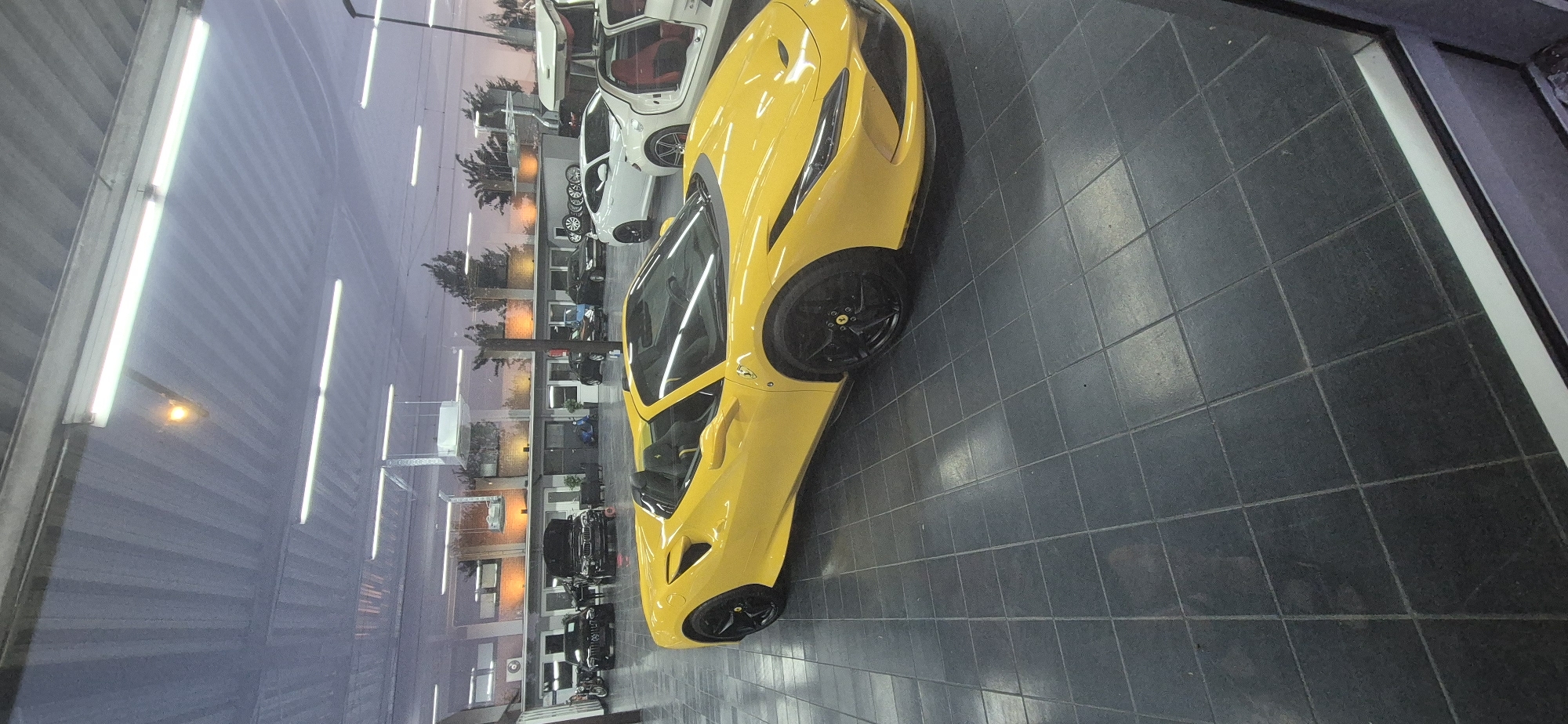 This image captures a yellow Ferrari parked indoors on a tiled floor. The car is the central subject, with its sleek lines and vibrant color prominently displayed. The setting appears to be a car showroom or a garage, indicated by the presence of other vehicles in the background, including a white luxury car with its doors open, revealing a red interior. The lighting suggests it might be evening or a dimly lit interior space, with fluorescent lights visible along the ceiling. Reflections on the floor and the cars indicate a glossy surface. The overall impression is one of luxury and automotive display. No people are visible in the frame. There is no visible text. The location context is Mainz, Germany, though this cannot be determined from the image itself.