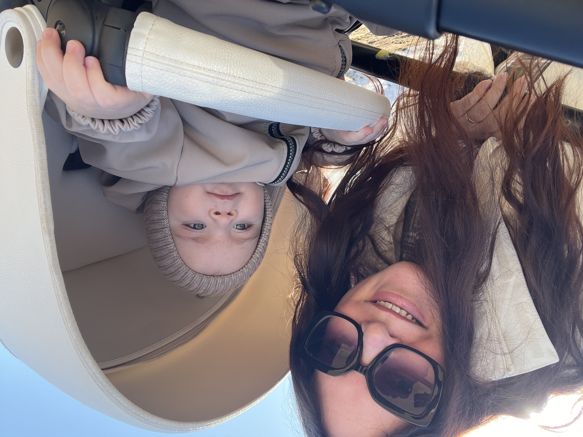 A low-angle, selfie-style photograph shows a woman and a young child, both positioned upside down from the perspective of the viewer. The woman has dark hair and is wearing sunglasses. She is smiling, and a ring is visible on her finger. The child, who is wearing a knitted cap and a light-colored hooded outfit, is looking directly at the camera. The two are in a stroller, which is visible surrounding them, with the blue sky in the background. The location is Noviy, Russia.