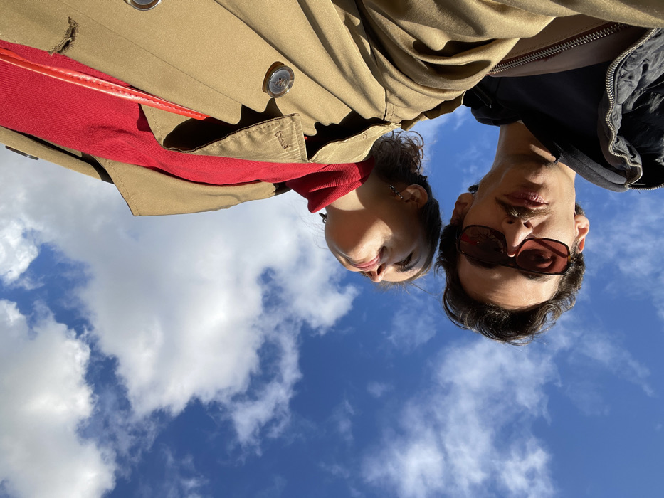 A low-angle outdoor photograph depicts two individuals, one female and one male, positioned against a clear blue sky featuring scattered white cumulus clouds, indicating a daytime scene in Budva, Montenegro.
The female subject, situated in the upper-right portion of the frame, is visible from the shoulders up. She wears a vibrant red garment underneath a tan trench coat, with a thin red strap, possibly from a bag, visible over her right shoulder. Her head is tilted backwards and slightly to her left, with her eyes directed upwards or closed. She exhibits dark hair and multiple earrings on her visible ear.
The male subject, positioned in the lower-left portion of the frame, is also visible from the shoulders up. He wears a dark, possibly black, hooded jacket and dark sunglasses with reddish-brown tinted lenses. He possesses dark hair and a mustache, with his face angled slightly upwards towards the camera.
Both subjects are illuminated by natural sunlight from above, casting distinct shadows. Their apparent interaction involves posing for the camera from a low vantage point. The background consists solely of the sky and clouds.