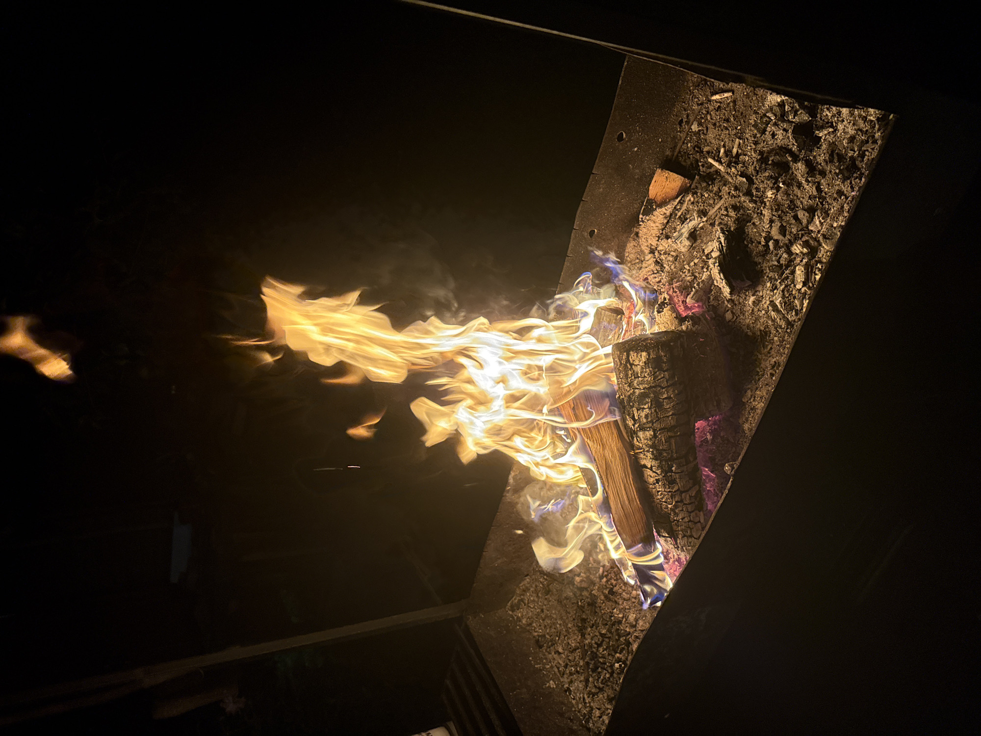 In the city of Автомобилист-1 Снт, Russia, a fire burns brightly within a dark, rectangular metal fire pit. The fire is composed of several logs, with vibrant orange and yellow flames reaching upwards, interspersed with wisps of white smoke. The ground around the fire is covered in ash and embers, with some larger pieces of charcoal visible. The surrounding area is dark, suggesting the photograph was taken at night. There are no people visible in the frame. The fire pit itself has a textured, dark interior. The overall scene depicts a contained outdoor fire, likely for warmth or cooking.
