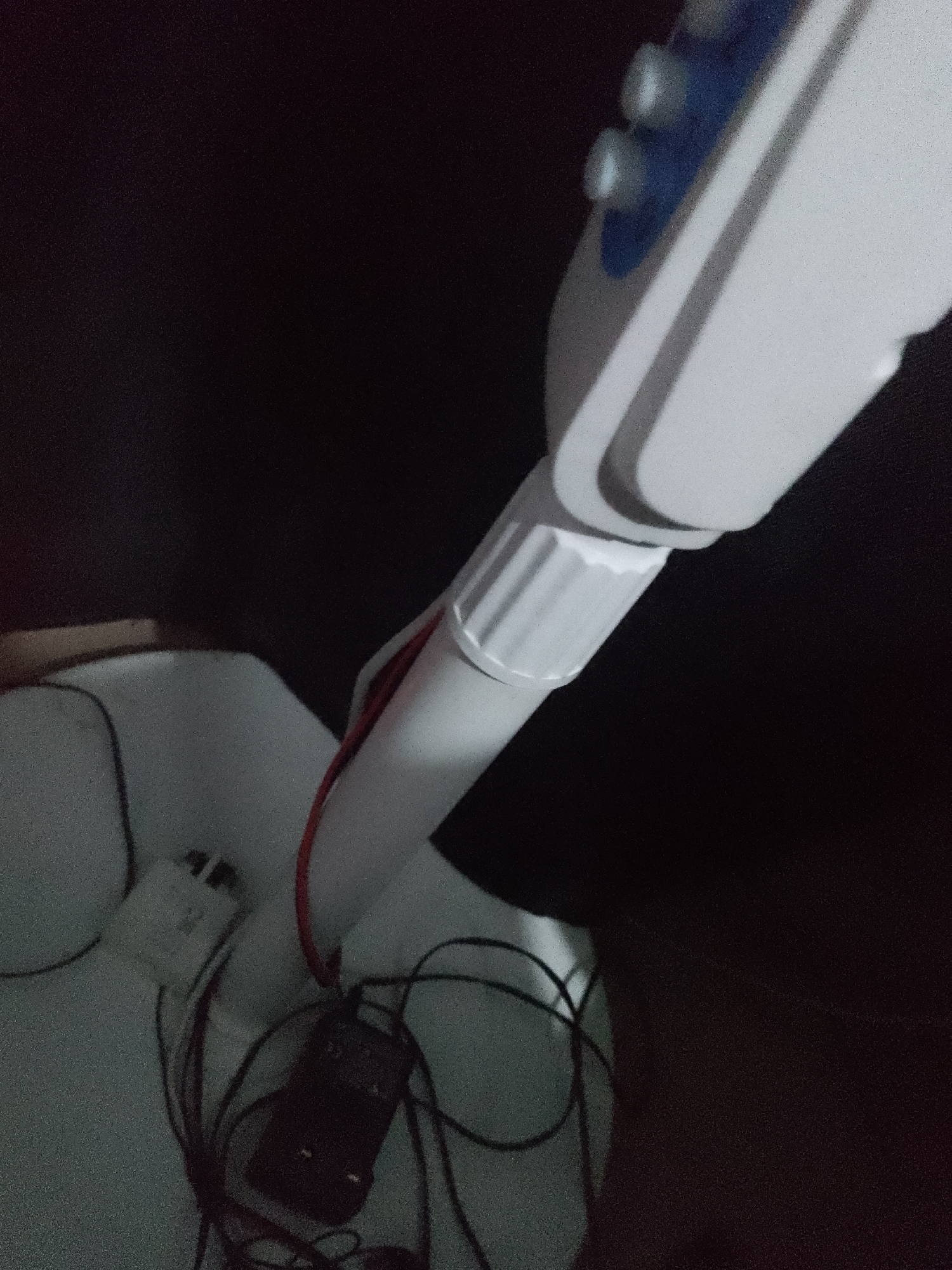 The image shows a close-up of a white and blue medical device, possibly a dental X-ray machine, with a dark background. The device has a long, articulated arm with a white, cylindrical extension that has ribbed detailing. Two blue buttons are visible on the upper part of the device. Red and black wires are connected to the extension of the arm and lead to some black electronic components. The background is predominantly black, suggesting a dimly lit environment. The scene is indoors, but the specific location within a city is unknown. There are no people visible in the image.