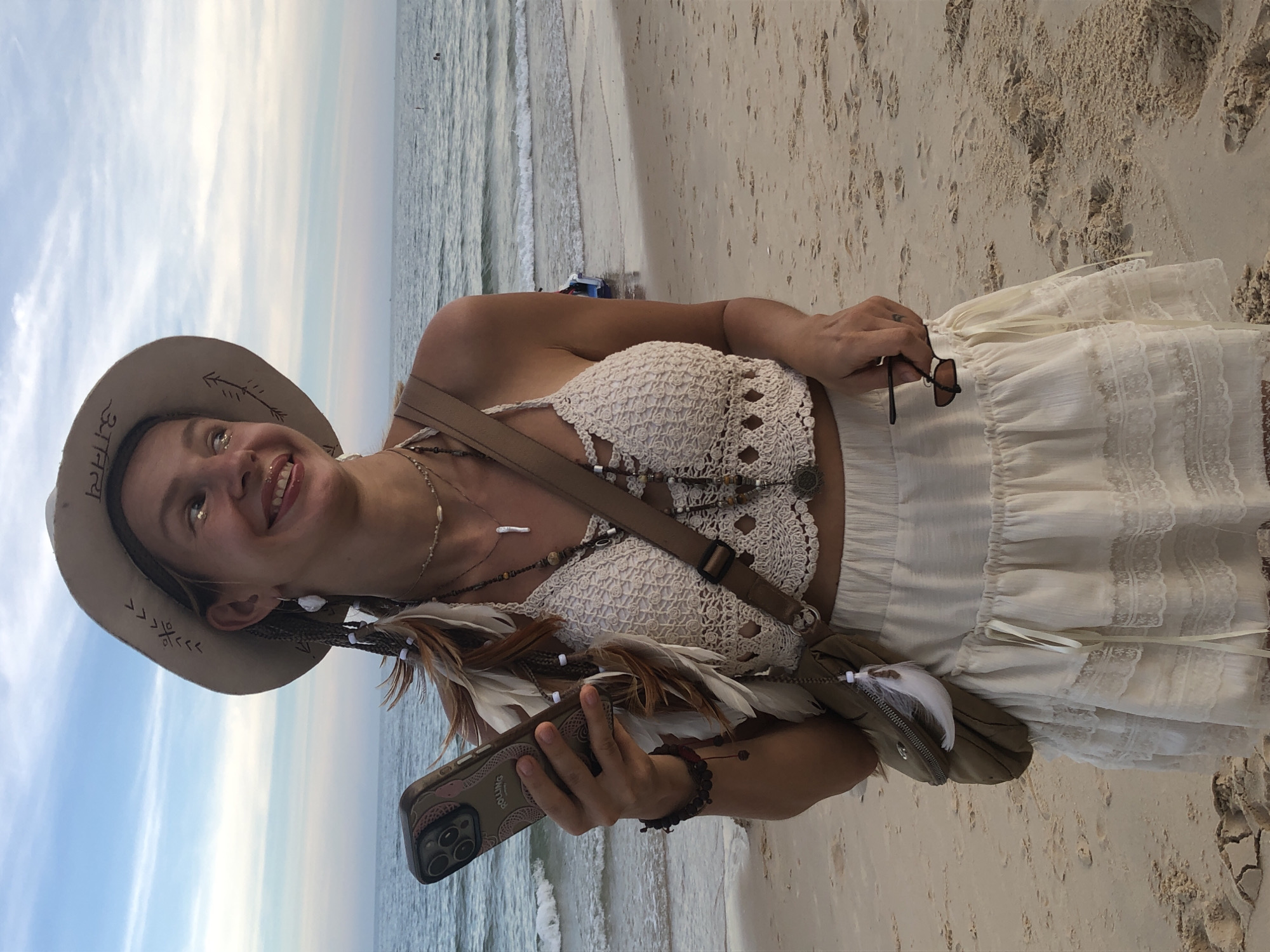 A young woman stands on a sandy beach, holding a smartphone in her left hand. She wears a white, crocheted halter top, a white, layered skirt with lace trim, and a wide-brimmed hat adorned with feathers and tribal markings. She has braided hair with feathers and beads woven into it. A crossbody bag is slung over her right shoulder. Her right hand holds a pair of sunglasses. The ocean is visible in the background with gentle waves and a boat in the distance. The sky is a soft blue with wispy clouds. The scene appears to be a coastal area in Phu Thuy Ward, Vietnam.