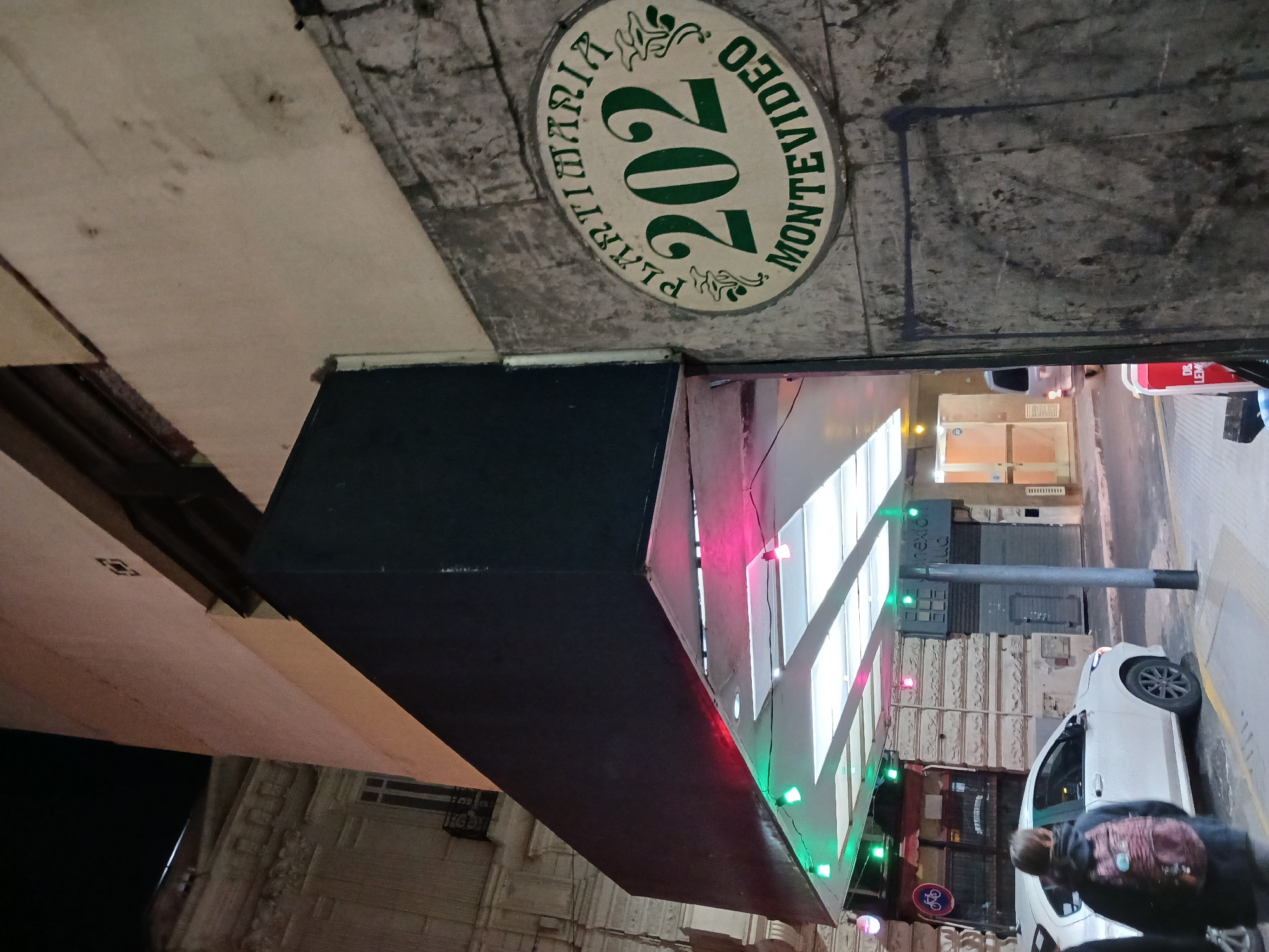 A nighttime street scene shows a white car parked along a sidewalk with a person wearing a dark jacket and backpack standing beside it. Above, a building overhangs the sidewalk, decorated with green and red lights. Painted on the building's facade is a circular sign reading "PLAYA TIMARIA 202 MONTEVIDEO" in green lettering on a white background. The location appears to be in Montevideo, as indicated by the sign.