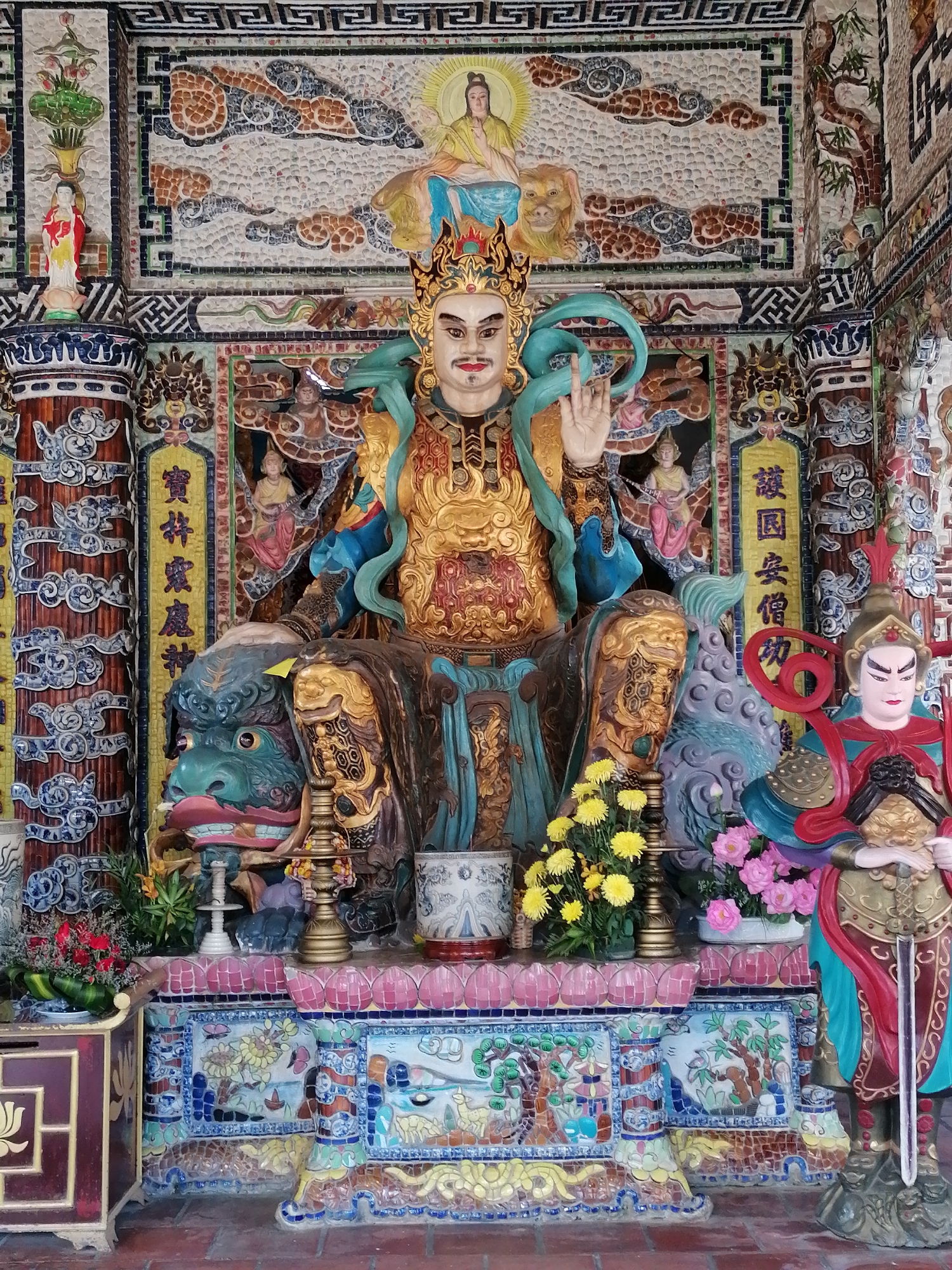 The image shows a large, ornate statue of a seated deity or warrior within what appears to be a temple or shrine. The figure is richly decorated with gold and blue attire, a crown, and is seated on a green mythological creature resembling a dragon or lion. The background is elaborately tiled with mosaic patterns featuring Buddhist or Taoist imagery, including figures, animals, and celestial elements. To the right of the main statue, a smaller, armored statue of a warrior stands guard, holding a sword. In front of the statues, there is a floral arrangement of yellow chrysanthemums and pink flowers, and a decorative altar with various ceramic vessels and figurines. The floor is made of red brick. The temple is located in the city of Xuân Thành, Vietnam.