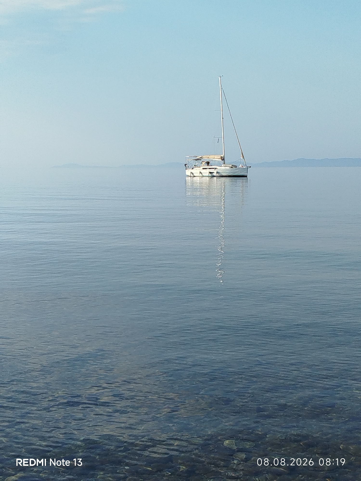 An overcast sky with a hazy horizon looms above a calm, blue sea in Sithonia Municipal Unit, Greece. The water is exceptionally still, with subtle ripples distorting the reflection of a single white sailboat. The sailboat, with its mast and sail furled, is positioned slightly off-center to the right. The seabed is visible through the clear, shallow water in the foreground, revealing a collection of rounded stones. The bottom left corner displays the text "REDMI Note 13", and the bottom right corner shows the date and time "08.08.2026 08:19". No people are visible in the frame.