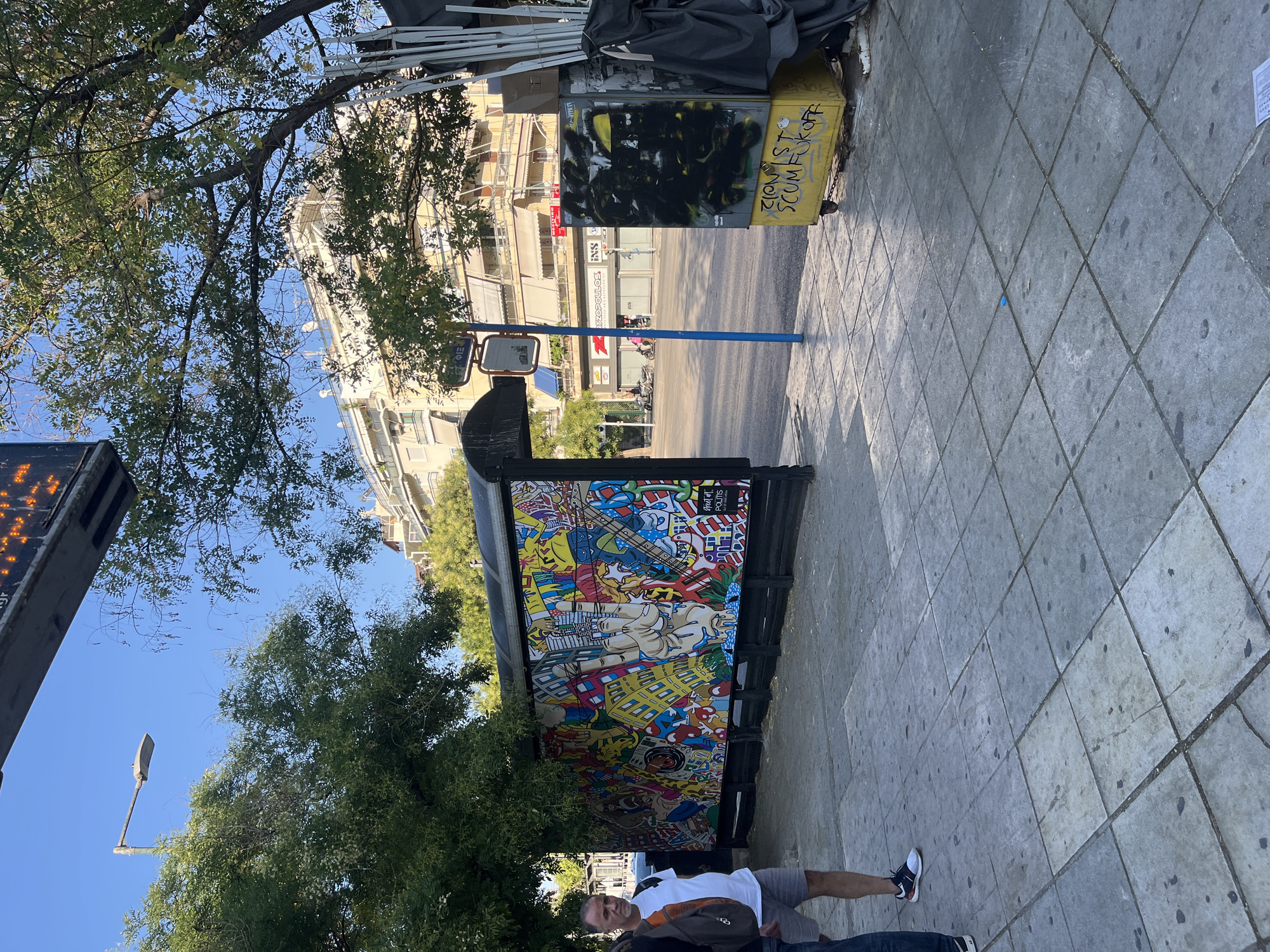 The image depicts a street scene in Athens, Greece. In the foreground, a sidewalk paved with square tiles leads to a bus stop shelter. The shelter's wall is adorned with a large, vibrant, and complex mural featuring cartoon-like imagery, cityscapes, and symbols such as a peace sign. To the right of the mural, a yellow waste bin with wheels is covered in black graffiti, and beneath it, yellow spray paint spells out "ZIONIST SCUMFUCKOFF." Two men are visible on the sidewalk, one in a white t-shirt and grey shorts, and another partially visible to his left wearing a backpack. Beyond the sidewalk and bus stop, a street runs parallel, with parked cars, motorcycles, and buildings lining the opposite side. One building prominently displays signage, including "RIZZOPOULOS" and "INN." A blue pole supporting a street sign and a mirror stands near the street. Trees with green foliage cast shadows on the sidewalk and street. The sky is clear and blue, indicating a sunny day.