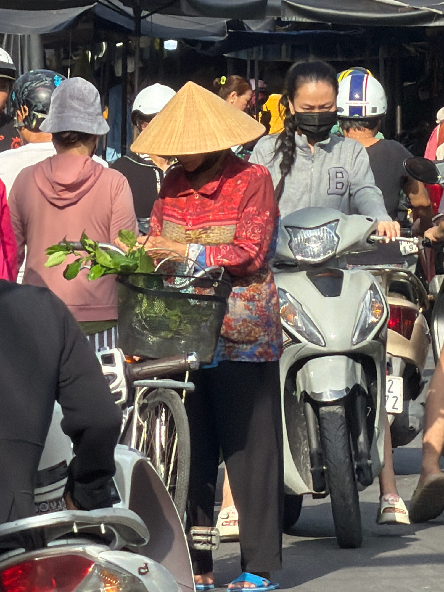 The image shows a street scene in Nha Trang, Vietnam, likely a market or a busy intersection. In the foreground, a person wearing a conical hat is holding a basket with green leaves. Next to them, another person in a gray jacket with a "B" emblem is riding a white scooter. Behind them, other individuals are visible, some wearing helmets. The environment appears to be outdoors with a makeshift canopy or awning overhead, suggesting a market stall or covered area. The overall impression is of a lively, everyday scene in a Vietnamese city.