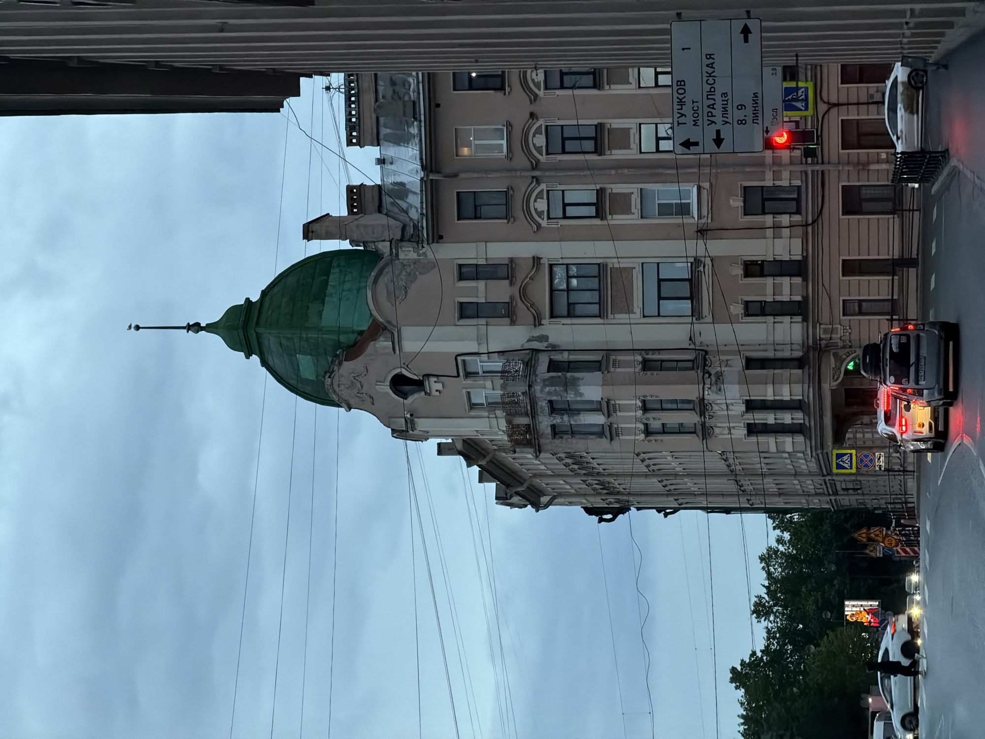 This image captures a street scene in Saint Petersburg, Russia, on a cloudy day. The focal point is a multi-story building with distinctive architecture, including a green domed roof topped with a spire and a small bird. The building features multiple windows, balconies, and ornate detailing. In the foreground, a street intersection is visible with cars, including a white sedan and a silver SUV, stopped at a red traffic light. A pedestrian is crossing the street. Traffic signs are present, indicating directions to "ТУЧКОВ МОСТ" (Tuchkov Bridge) and "УРАЛЬСКАЯ УЛИЦА" (Uralskaya Street). Overhead power lines crisscross the sky. Trees line the background to the left of the building. The overall atmosphere is subdued due to the overcast sky, suggesting either early morning or late evening.