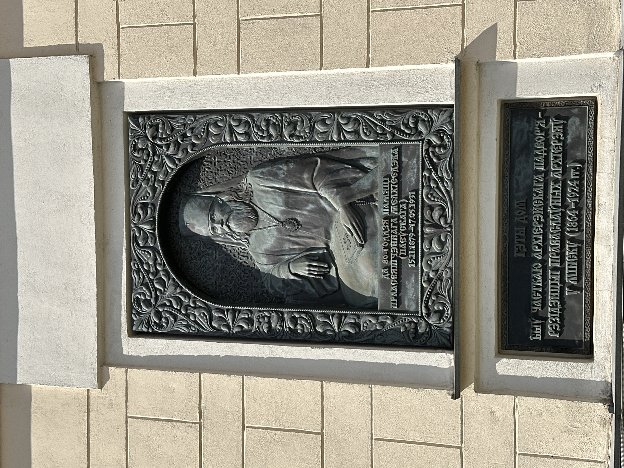 The image shows a close-up, eye-level, outdoor shot of a bronze commemorative plaque mounted on a textured, light-colored building wall. The plaque is rectangular with an ornate, embossed border featuring floral and scroll motifs. In the center of the plaque is a bas-relief portrait of an elderly man with a long white beard, wearing a religious headdress and ornate clerical robes. He is depicted seated, with one hand resting on an open book, and the other in his lap. Below the portrait are inscriptions in a Cyrillic script, providing biographical details and dates. Underneath this main plaque, there is a second, smaller rectangular plaque with further inscriptions, also in Cyrillic. The building wall appears to be made of large, rectangular stone blocks. The lighting suggests a sunny day, with shadows cast on the wall. The location is identified as the "House of Metropolitan Meletius Charnetsky" (Дом митрополита Милетия Чарнецкого) in Minsk, Belarus.
