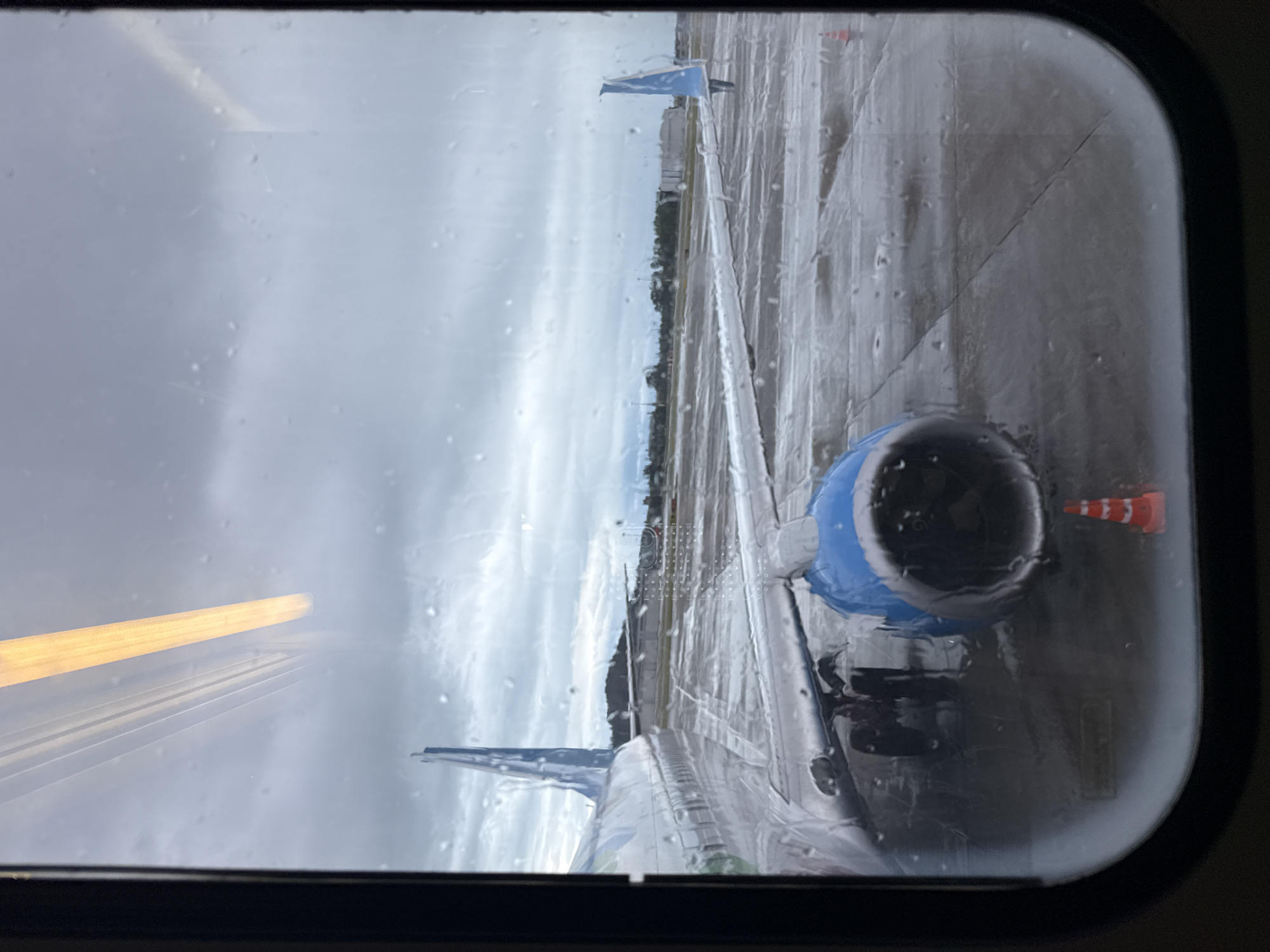 The image is a view from an airplane window. Rain obscures the glass. The airplane wing is visible, along with its engine, which has a blue cowling. The runway is wet. In the distance, there are buildings and trees under an overcast sky. The location is unknown, and it's NOT possible to determine if it's in Sochi, Russia, from the visual information provided.