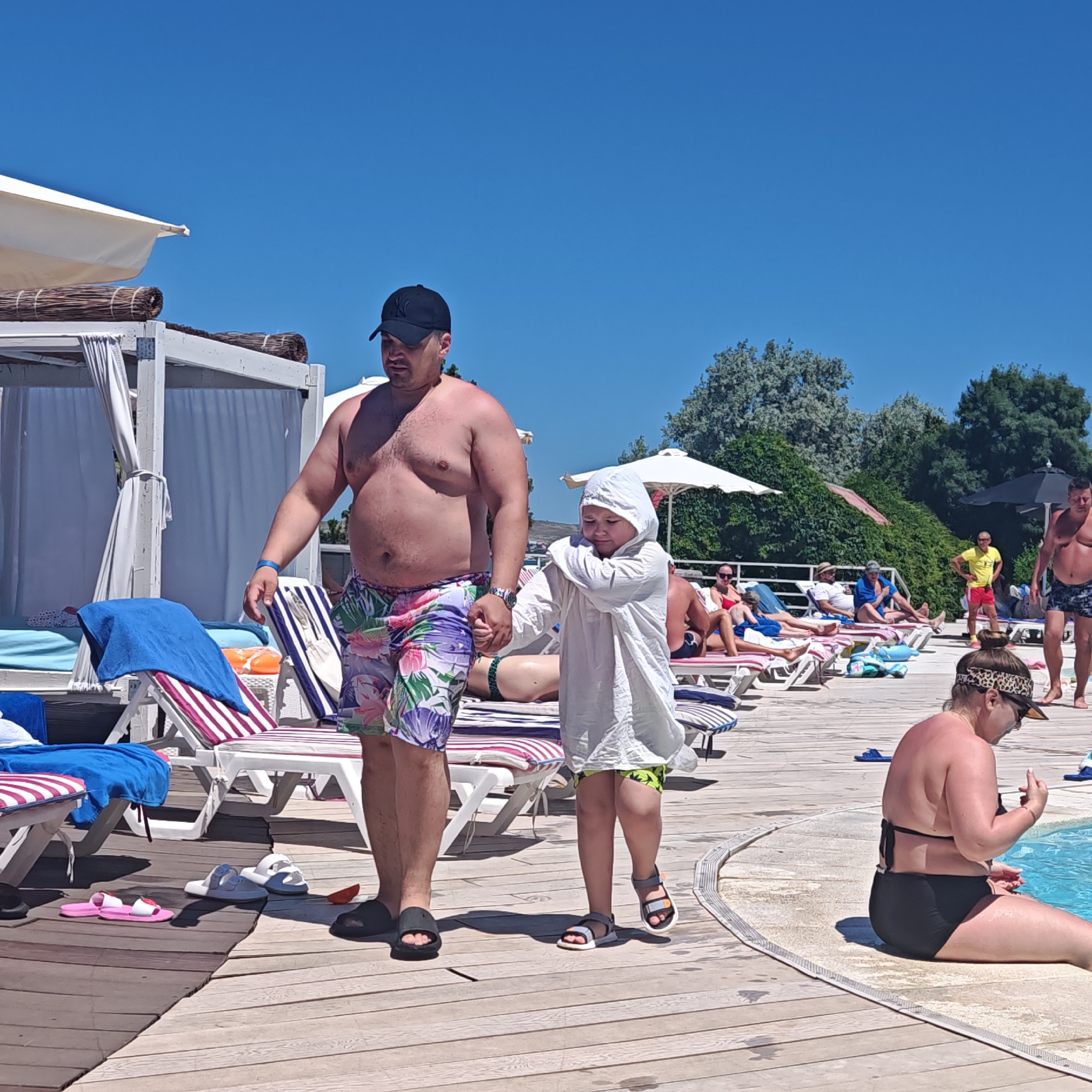 A man and a child are walking on a wooden deck. The man, wearing a black baseball cap and floral swim trunks, is holding the child's hand. The child is wearing a white hooded cover-up and green shorts. Behind them, several lounge chairs are set up, some with towels and other items. In the background, there are trees and buildings, and a clear blue sky. A woman in a black bikini is sitting by a pool in the lower right corner of the image. The scene appears to be a resort or a public pool area. The location is identified as Shcholkine, Ukraine.